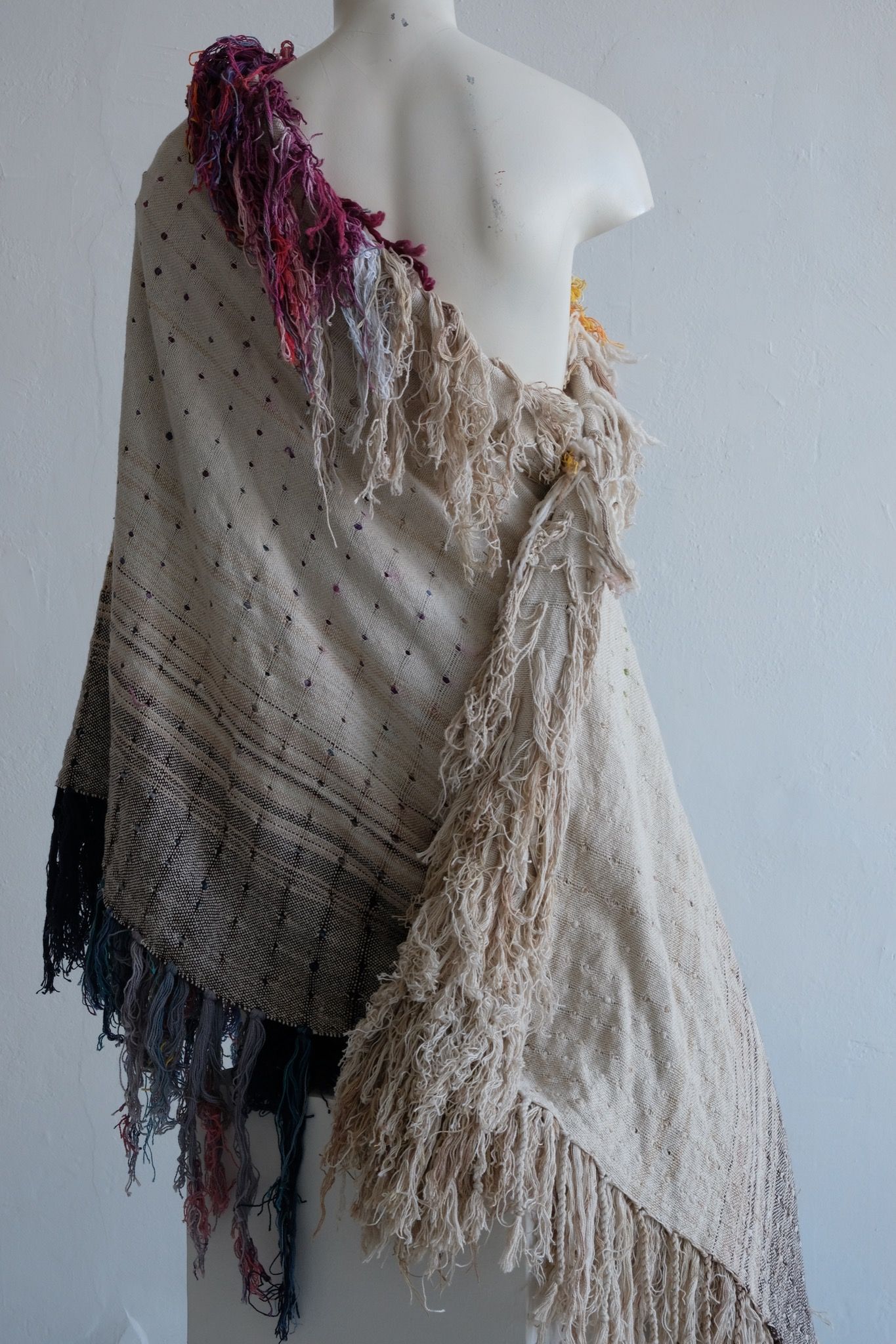 A rainbow, white and brown colored shawl, completely covered in fringe on a white mannequin, on a white pedestal, in a white room