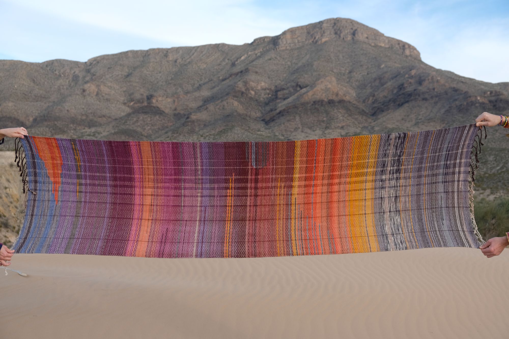 a handwoven brightly colored orange, yellow, purple, grey, blue shawl is held out by hands in front of a sand dune, desert mountain landscape