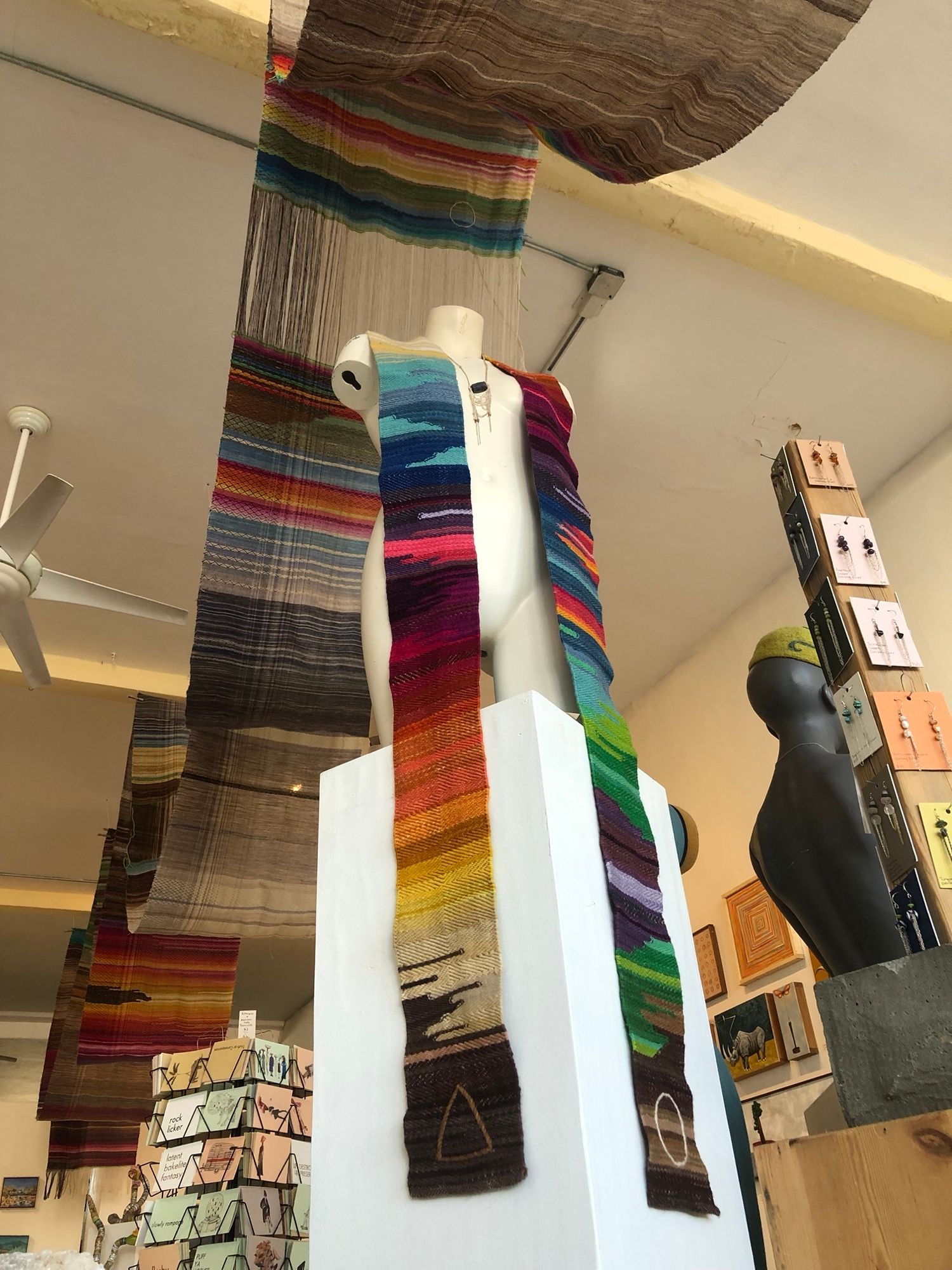 A white mannequin wearing a rainbow colored stole, A gallery space with white walls with paintings hanging, as well as brightly colored fabric draped, hanging from the ceiling 