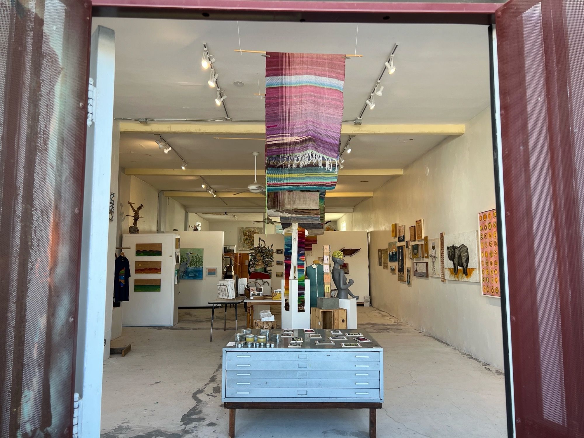 A gallery space with white walls with paintings hanging, as well as brightly colored fabric draped, hanging from the ceiling 