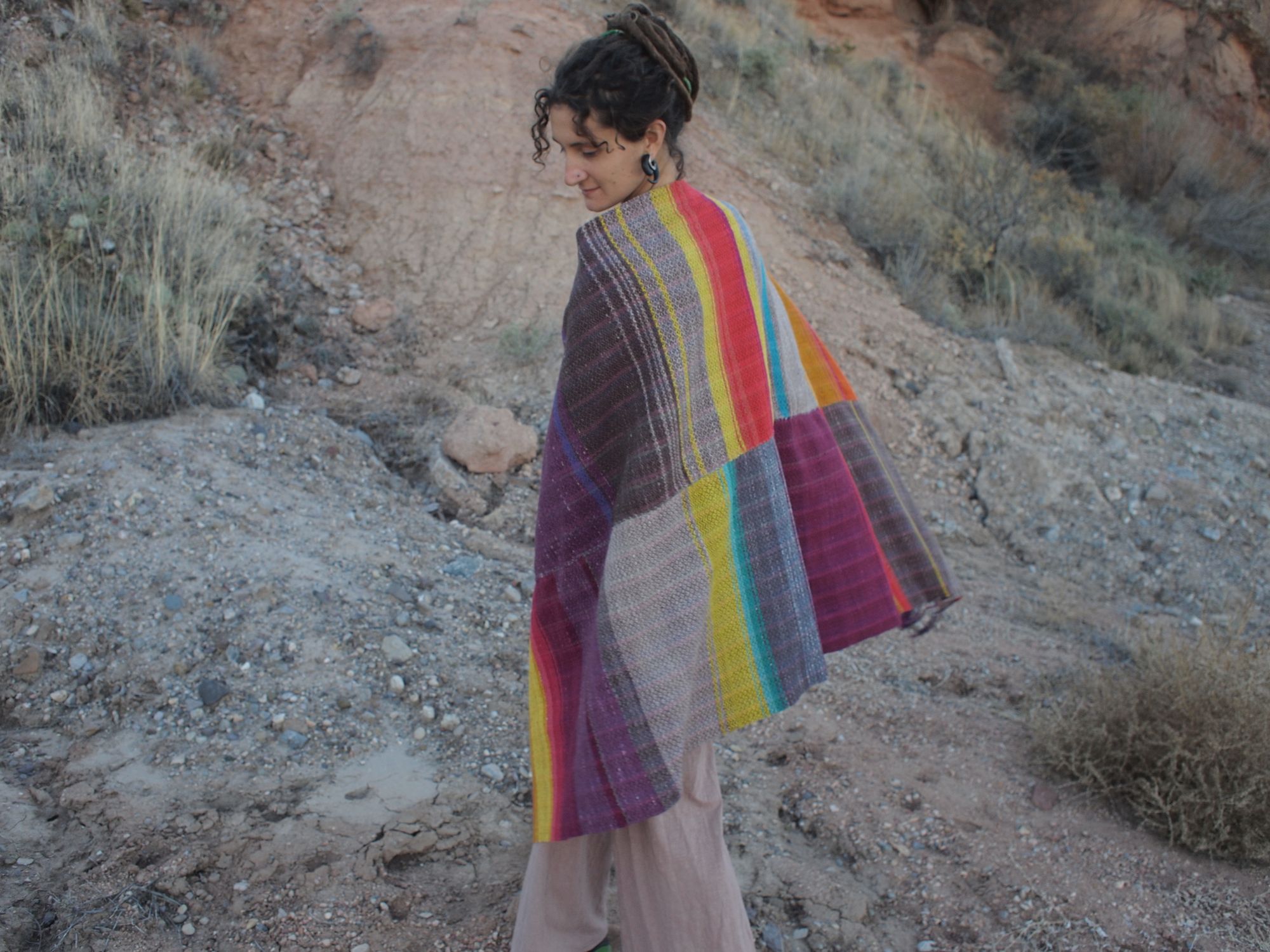 Woman in the desert, standing in dirt, wearing a blanket that is brown, grey, red, yellow, purple and blue