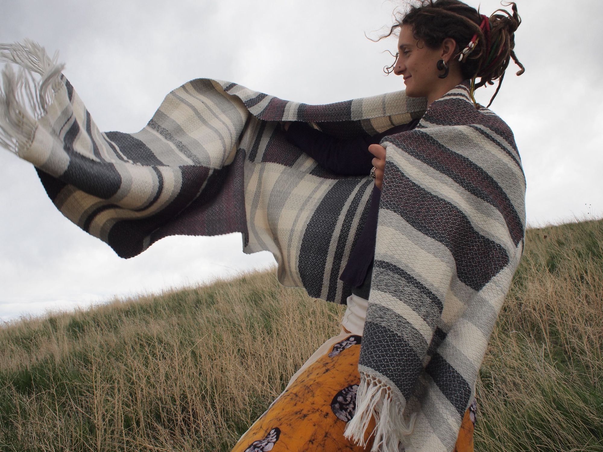 Woman wearing a dark grey, maroon and white blanket standing on a green grassy hill. The blanket is blowing in the wind. 