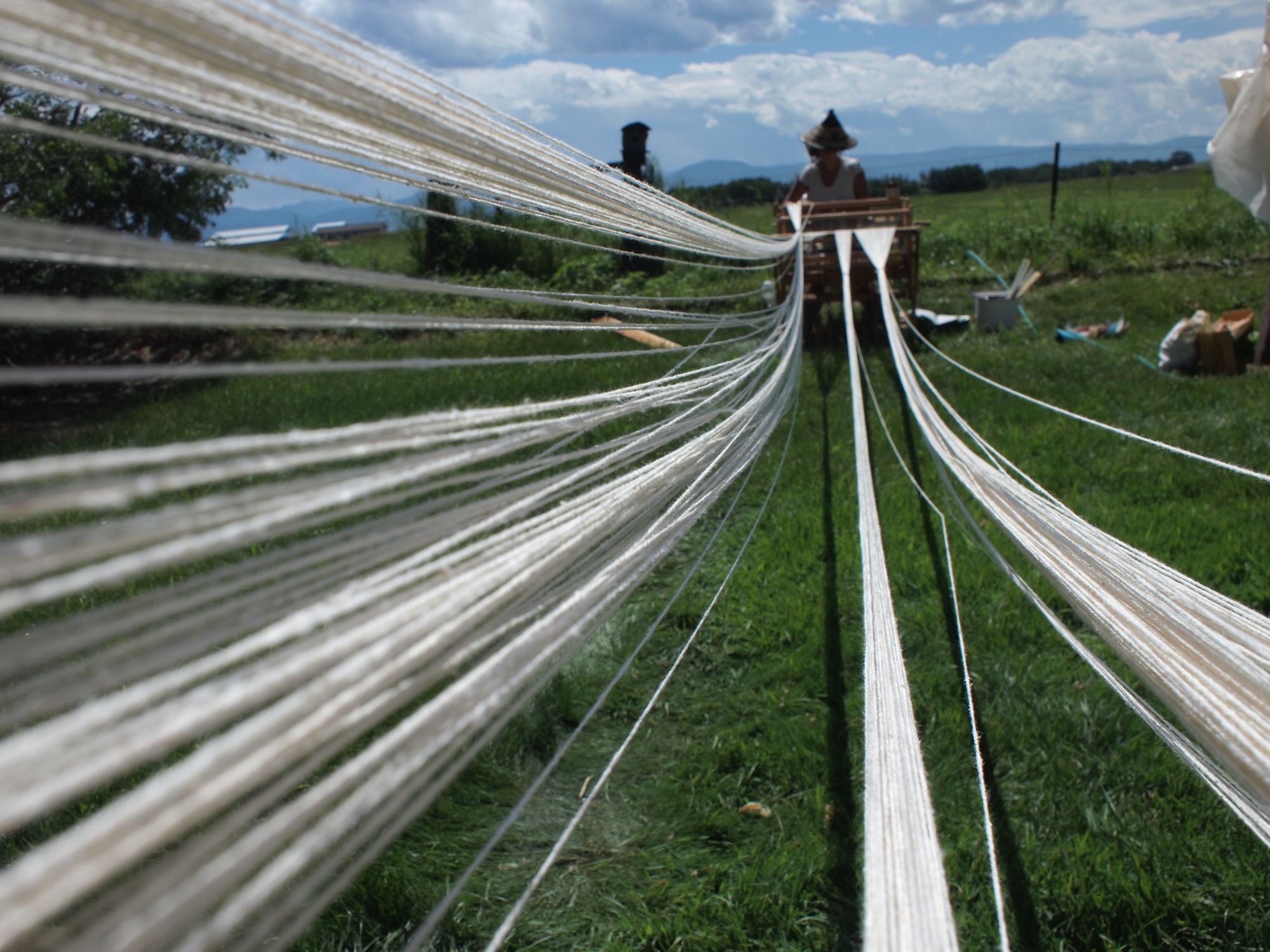 white strings lead from the camera to a woman in a pointy hat sitting at a loom in a green grassy field with blue cloudy skies and mountains in the background. 