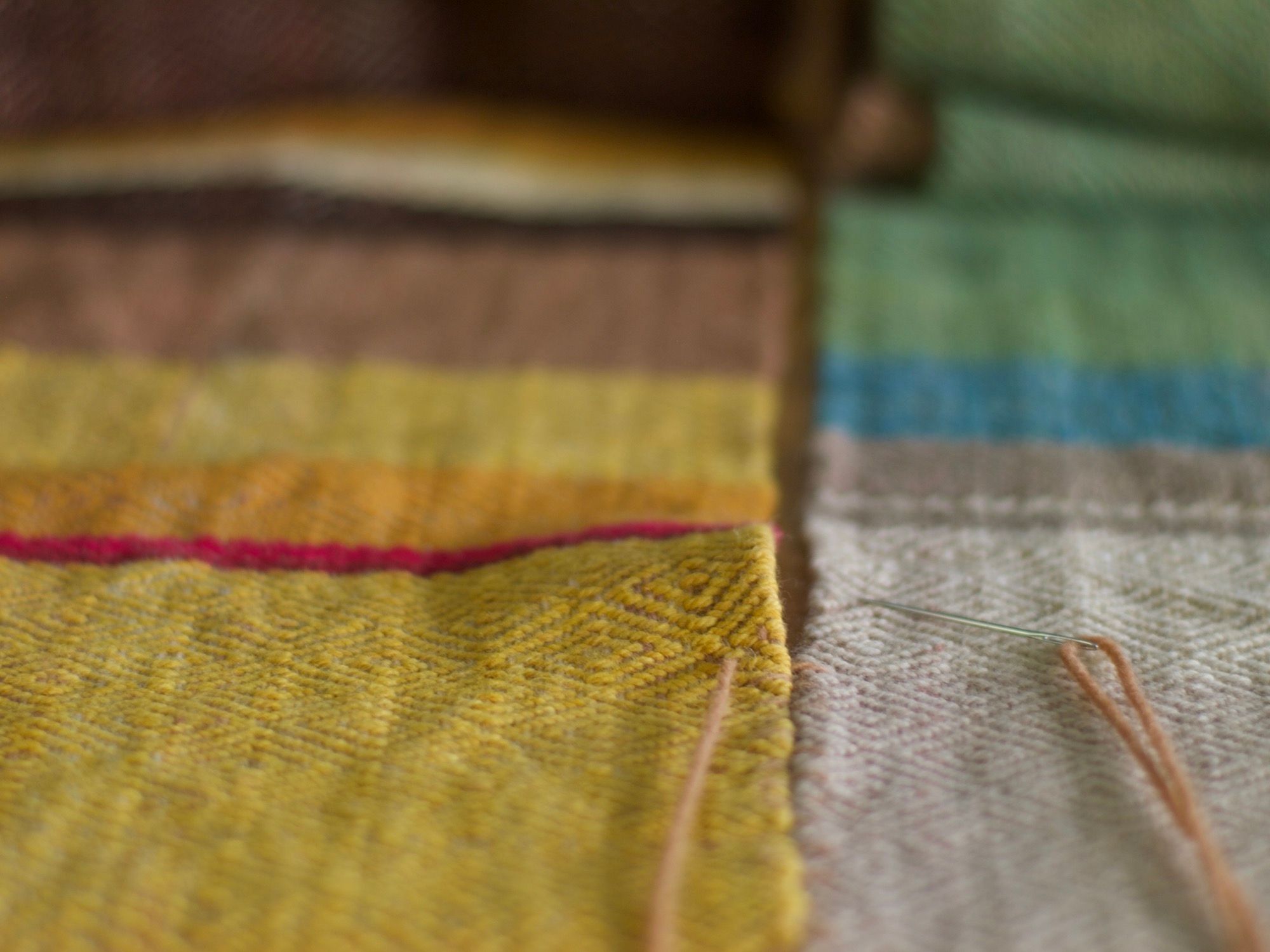 a needle sewing the panels of Details of a brown, pink, yellow, blue, green, tan and purple woolen blanket with a diamond pattern.