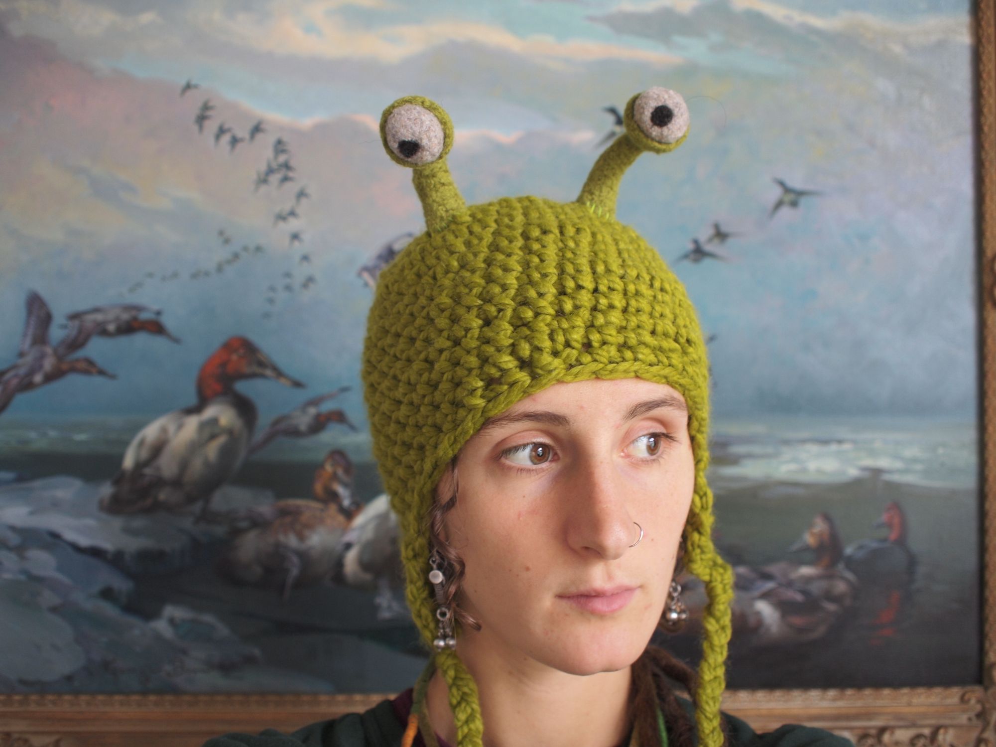 Woman wears a green hat with earflaps and stalked eyeballs in front of a painting with ducks.