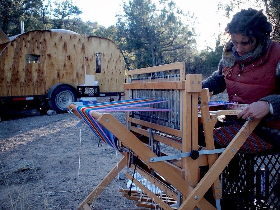 A woman in red puffy vest sits at a loom in the forest in front of a wooden teardrop camper.