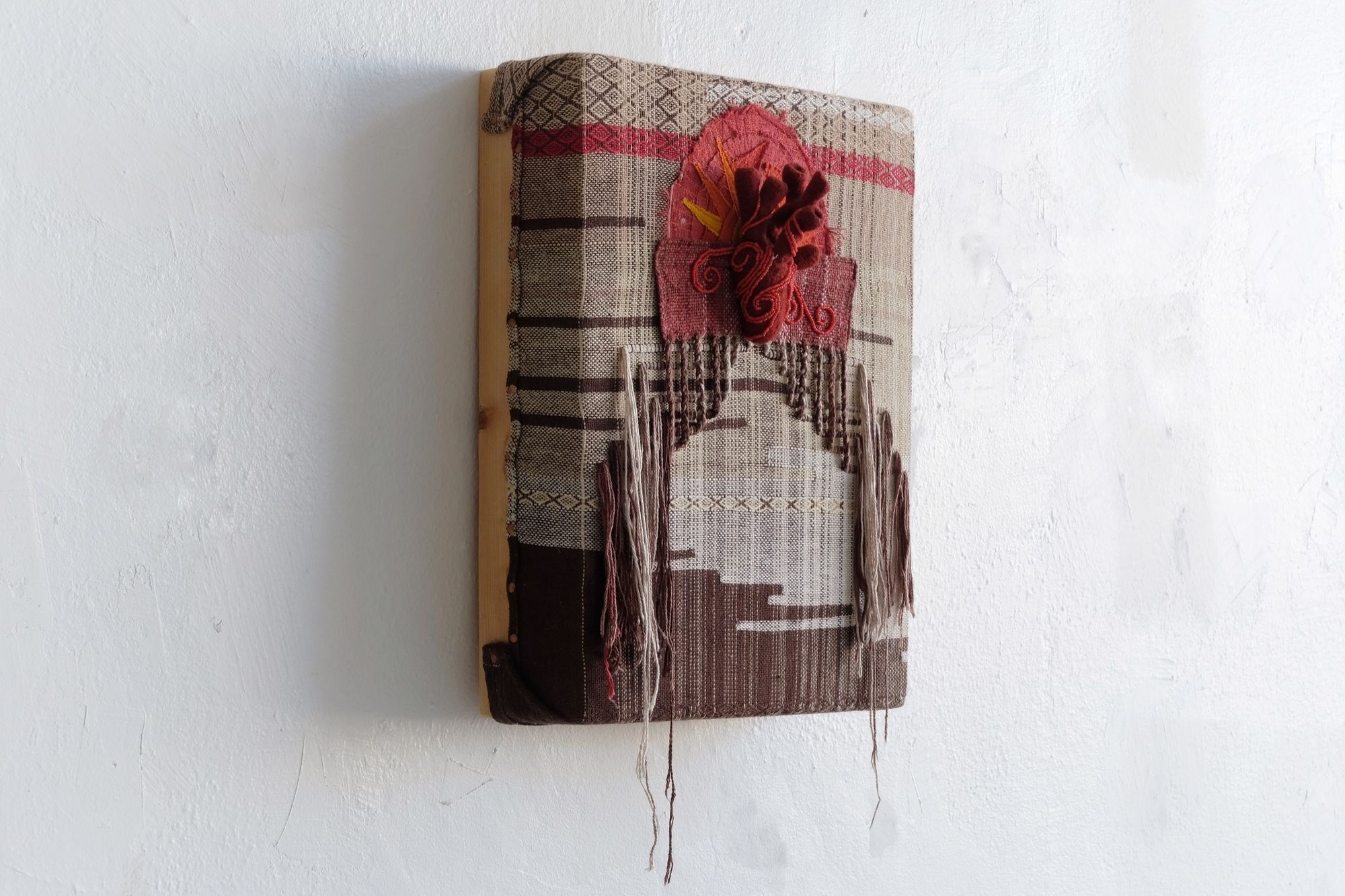 A textile wall sculpture with a red felted and beaded heart on textured handwoven brown, red and cream fabric. 