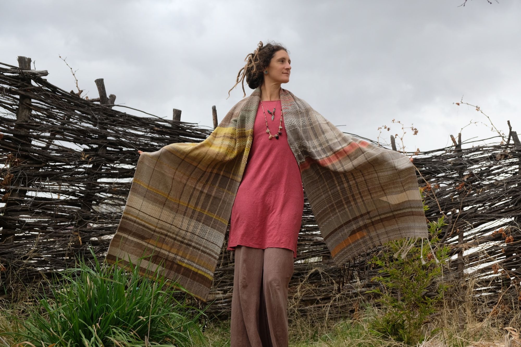 woman wearing a red-pink long-sleeved dress and a handwoven shawl against a stick fence