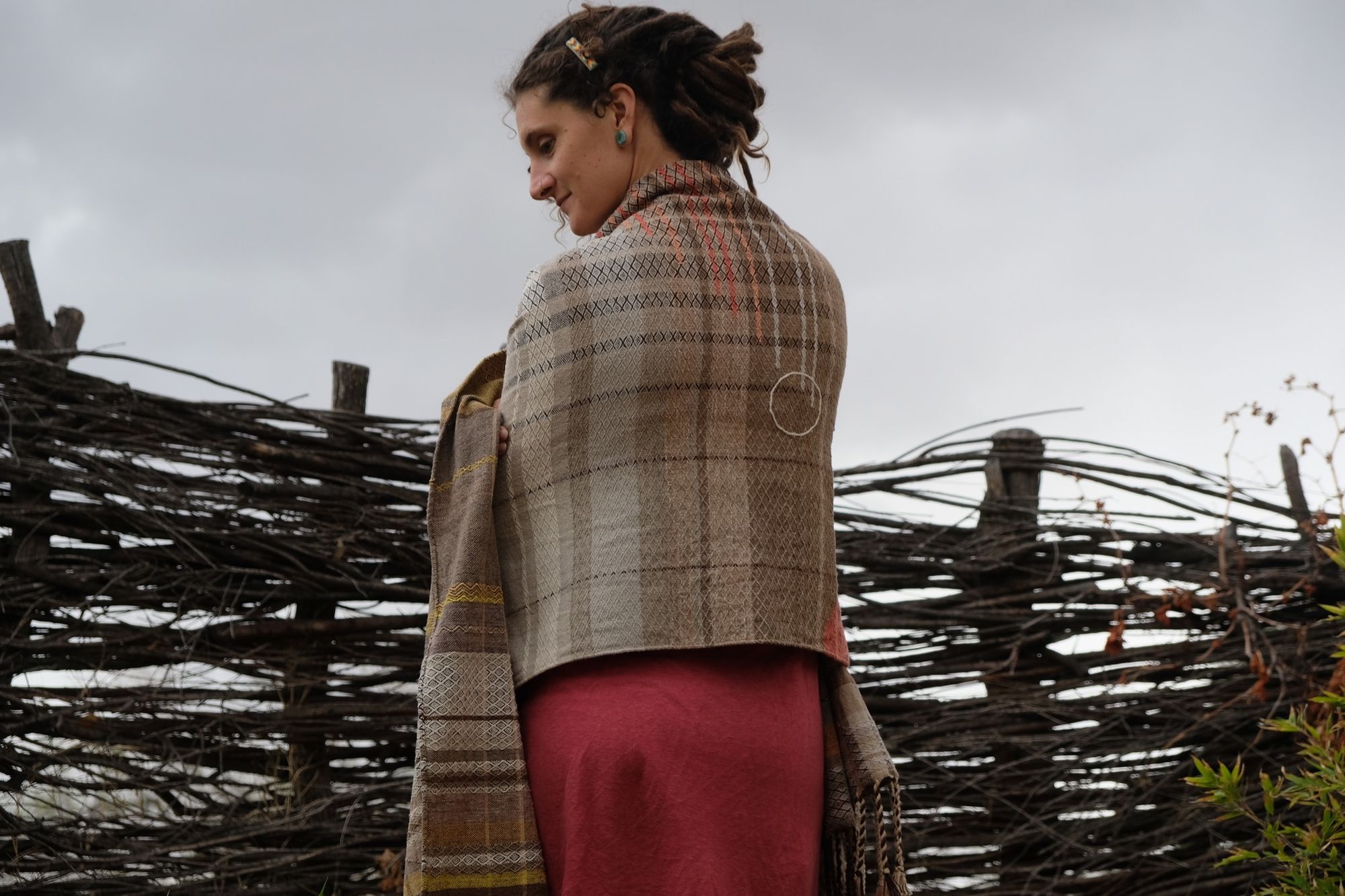 woman wearing a red-pink long-sleeved dress and a handwoven shawl against a stick fence