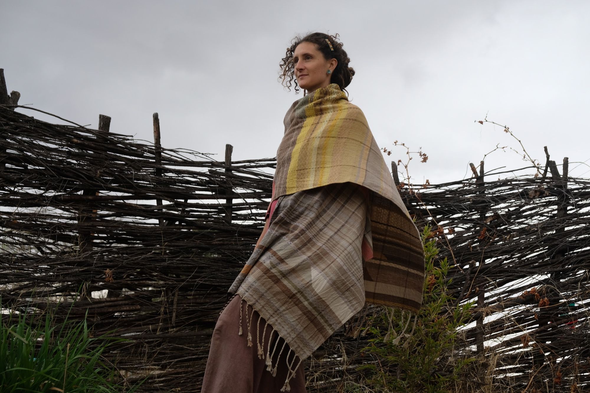 woman wearing a red-pink long-sleeved dress and a handwoven shawl against a stick fence