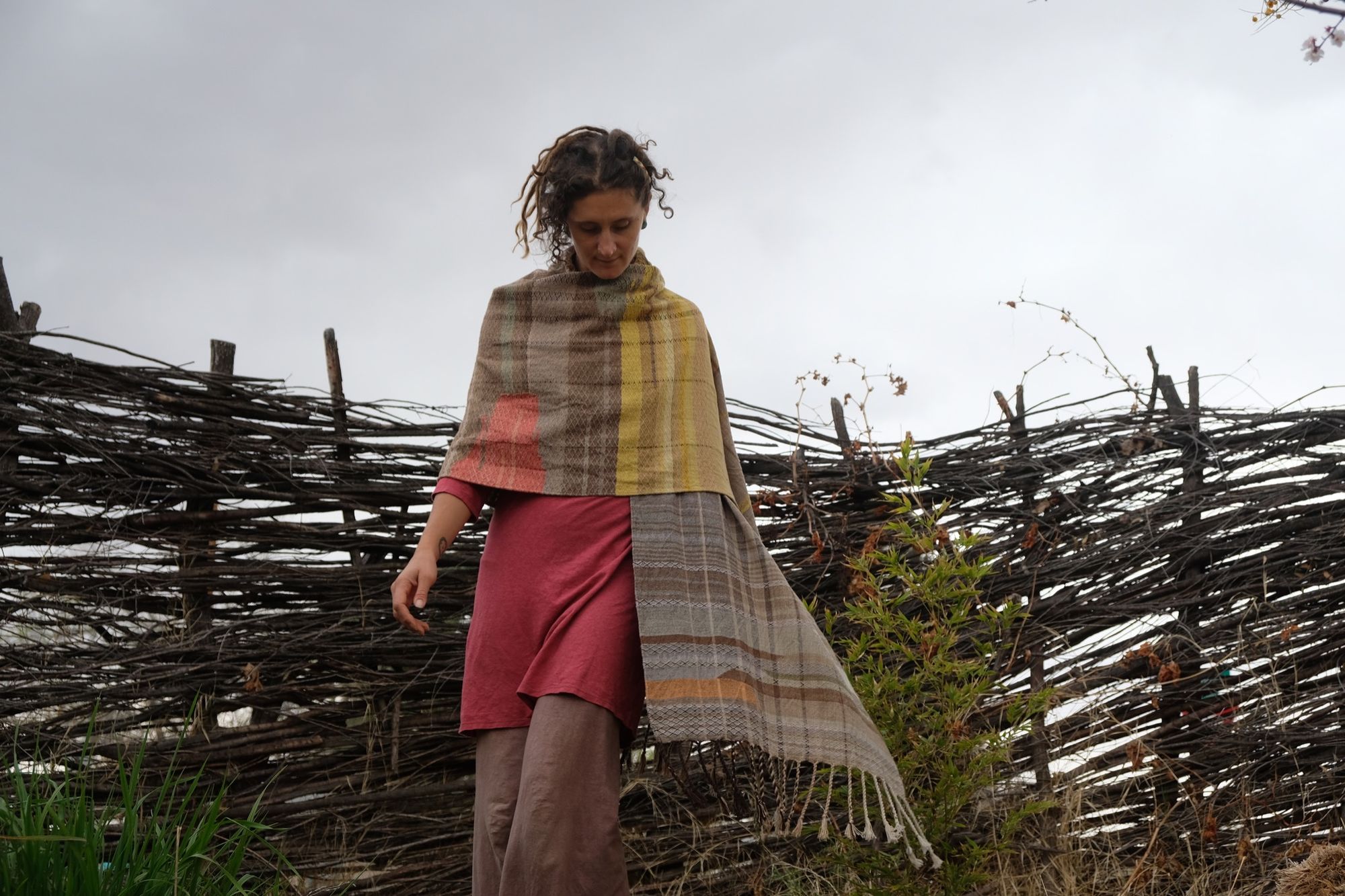 woman wearing a red-pink long-sleeved dress and a handwoven shawl against a stick fence