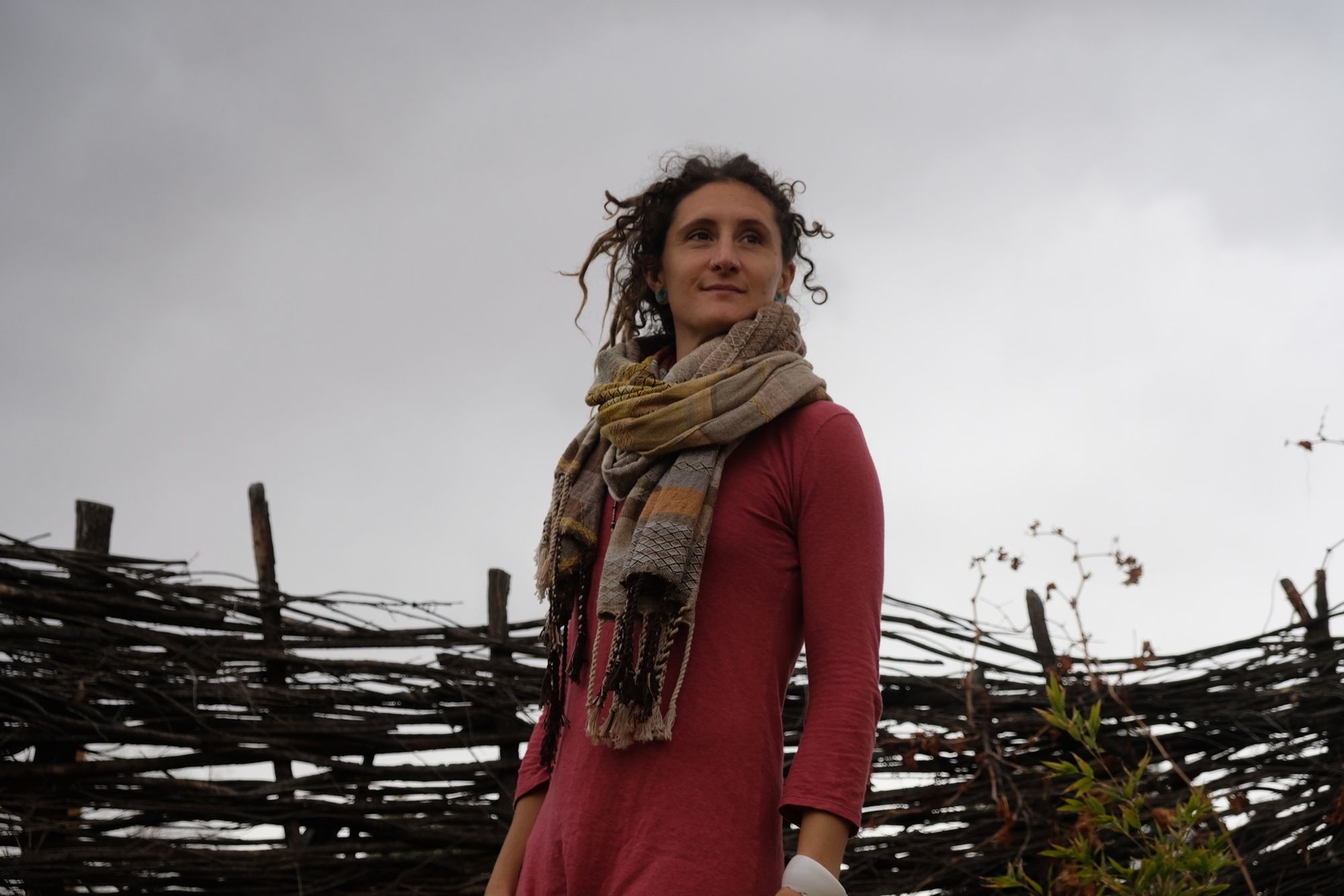 woman wearing a red-pink long-sleeved dress and a handwoven shawl against a stick fence