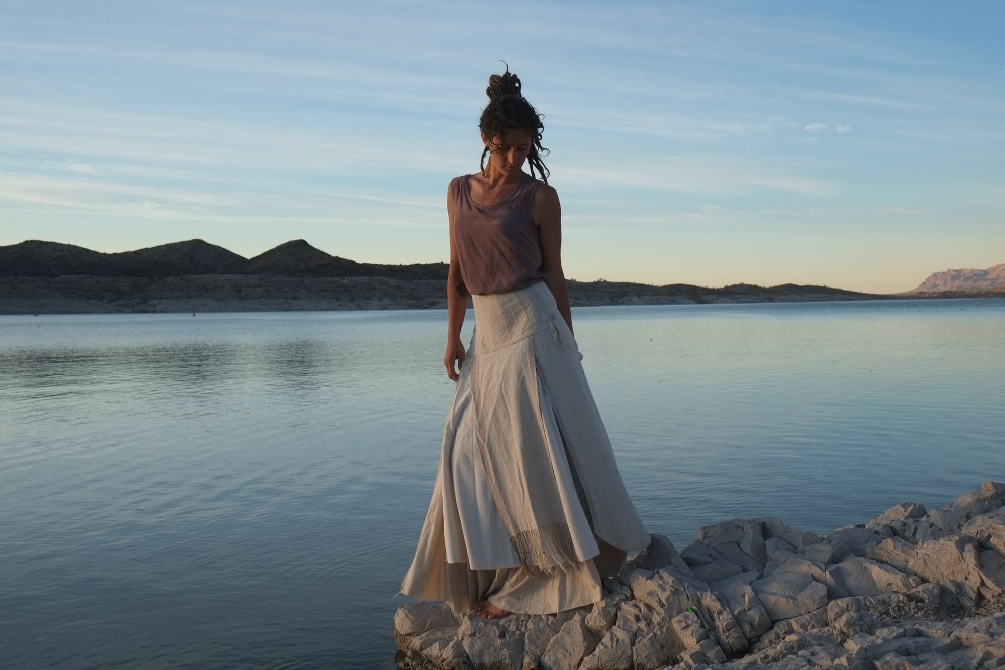 woman wearing white wedding skirt and lilac shirt by a still lake at sunset