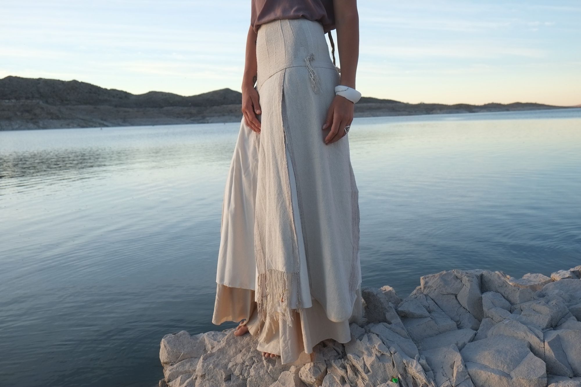 woman wearing white wedding skirt and lilac shirt by a still lake at sunset