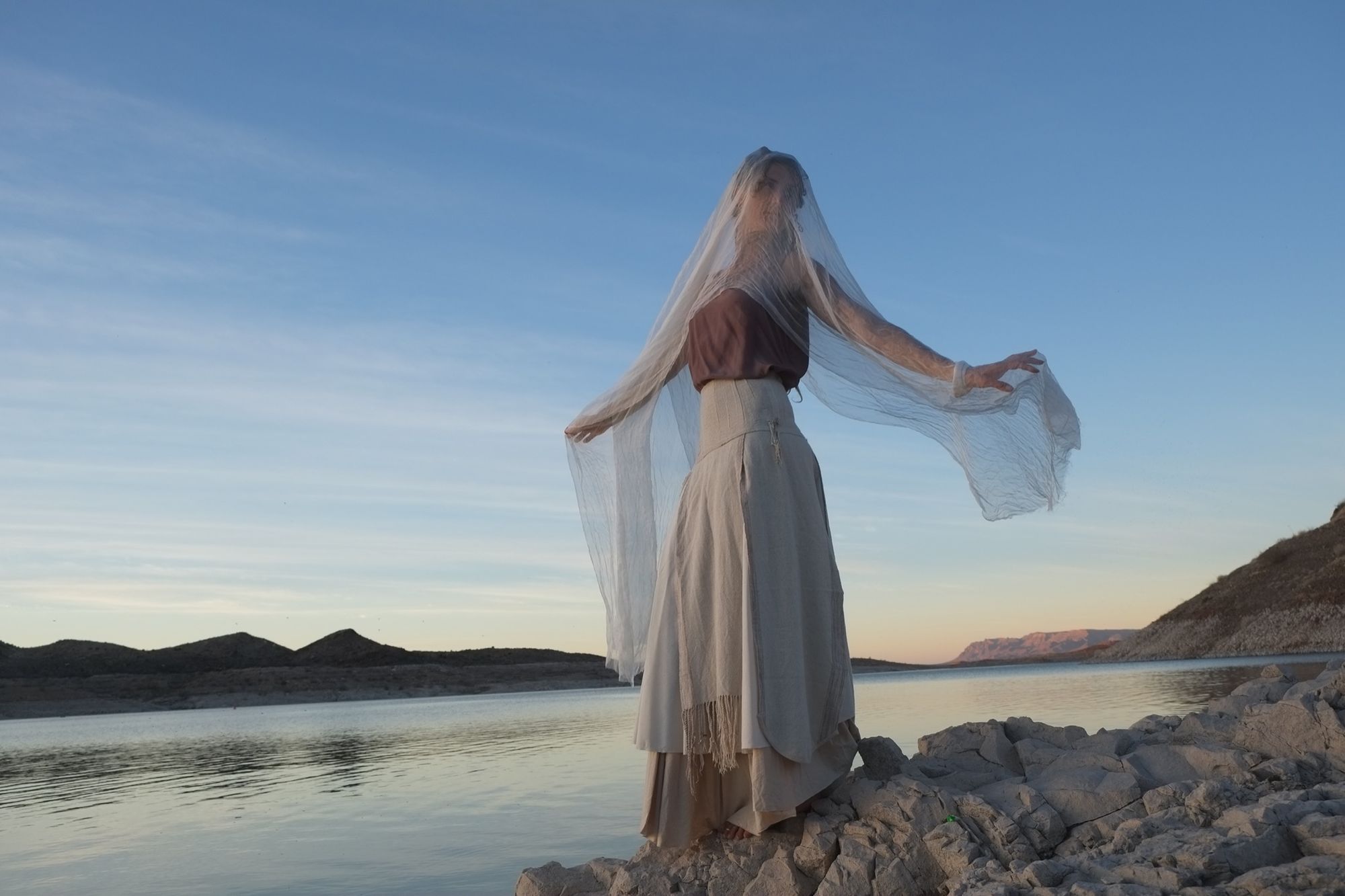 woman wearing white wedding skirt and lilac shirt by a still lake at sunset