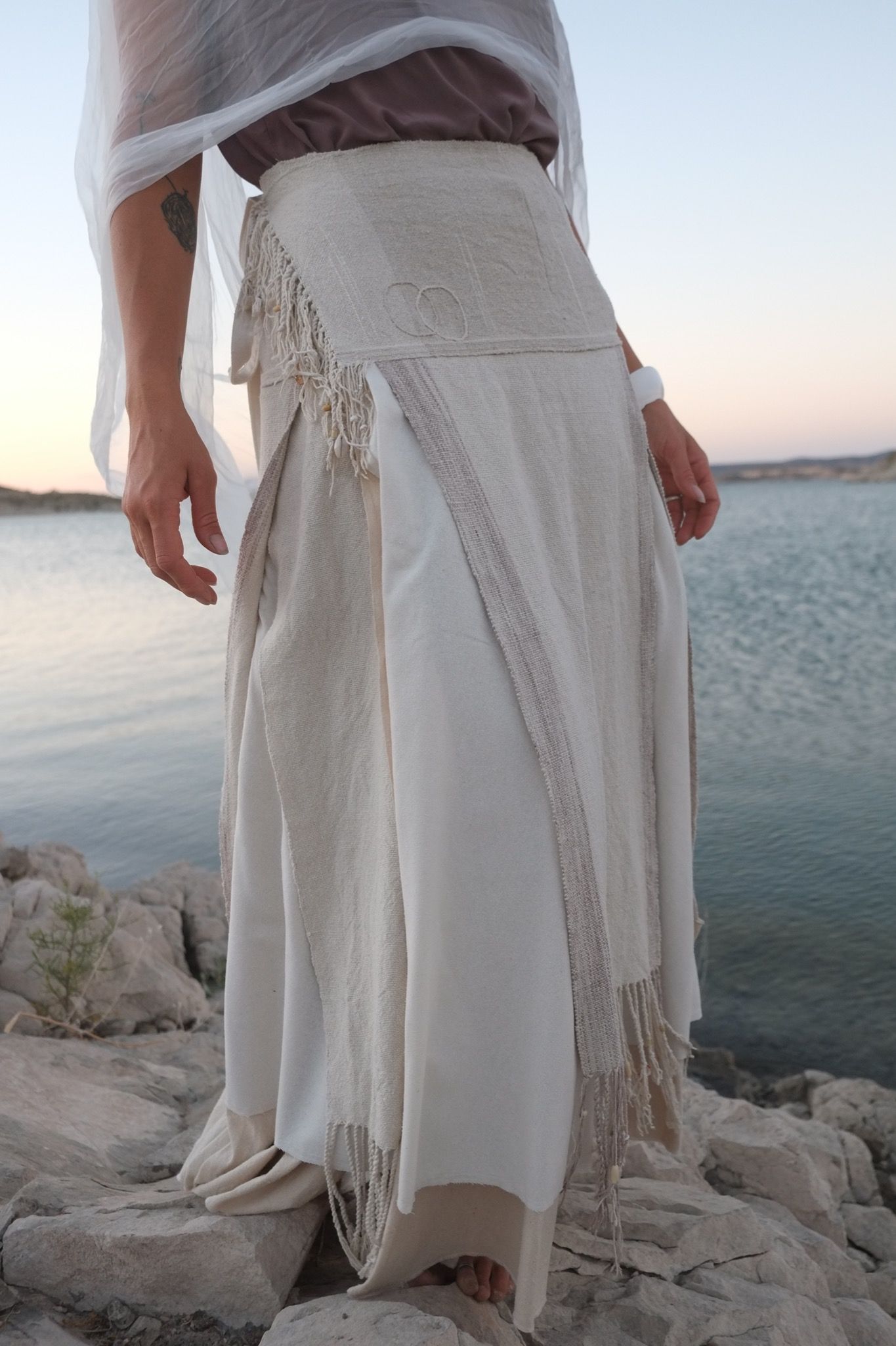 woman wearing white wedding skirt and lilac shirt by a still lake at sunset