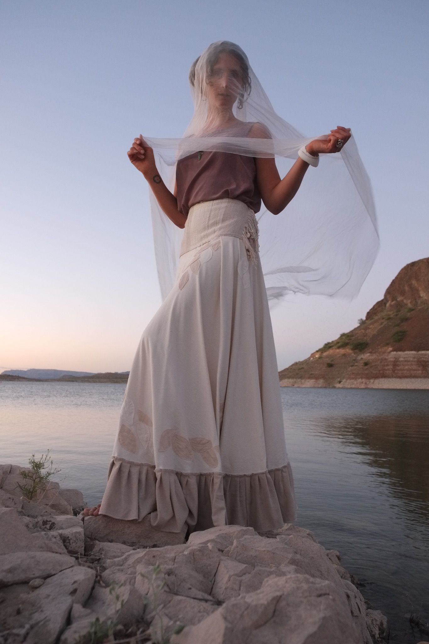 woman wearing white wedding skirt with thin veil by a still lake at sunset