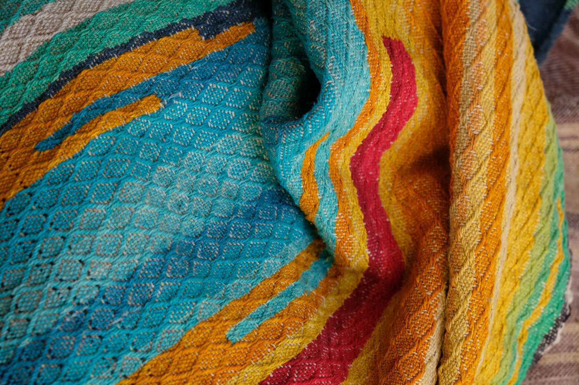 Handwoven fabric laying on a wooden floor, it is red, orange, blue, yellow, purple, brown and green