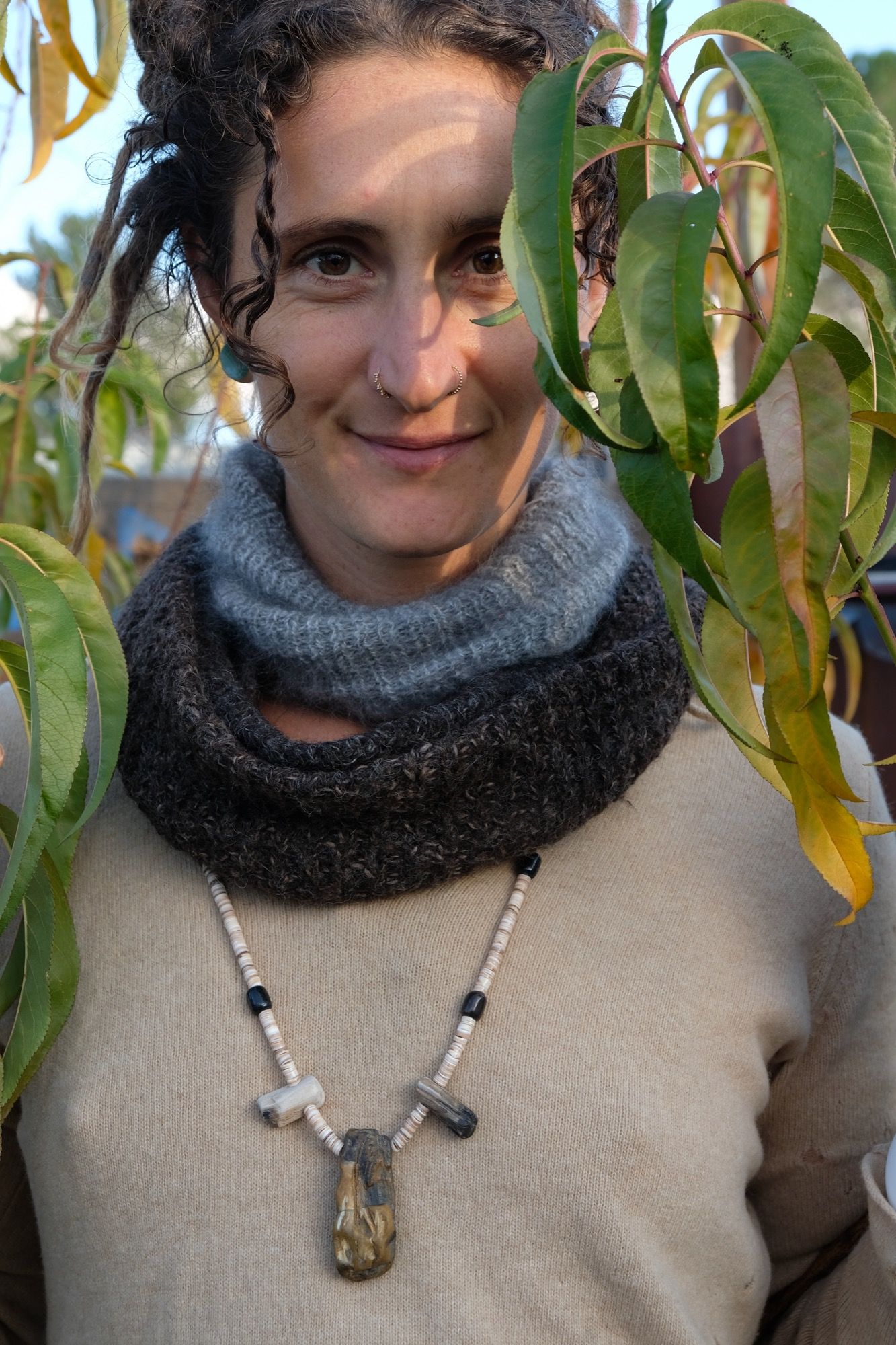 woman wearing tan sweater with a brown and grey fuzzy cowls and stone and shell necklace stands among the green and yellow leaves of a peach tree