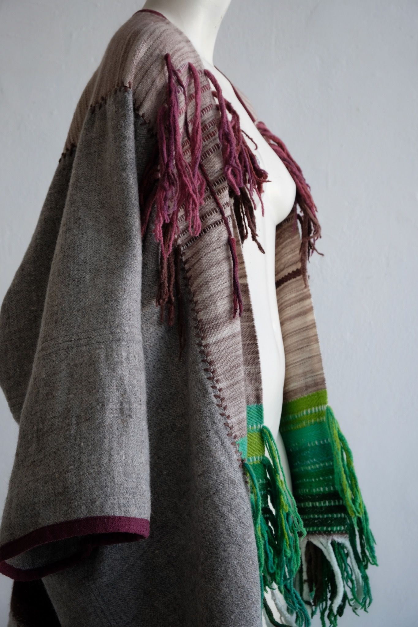 Handwoven, highly textured with fringe cloak that is grey, brown, green and rainbow on a white mannequin against a white wall