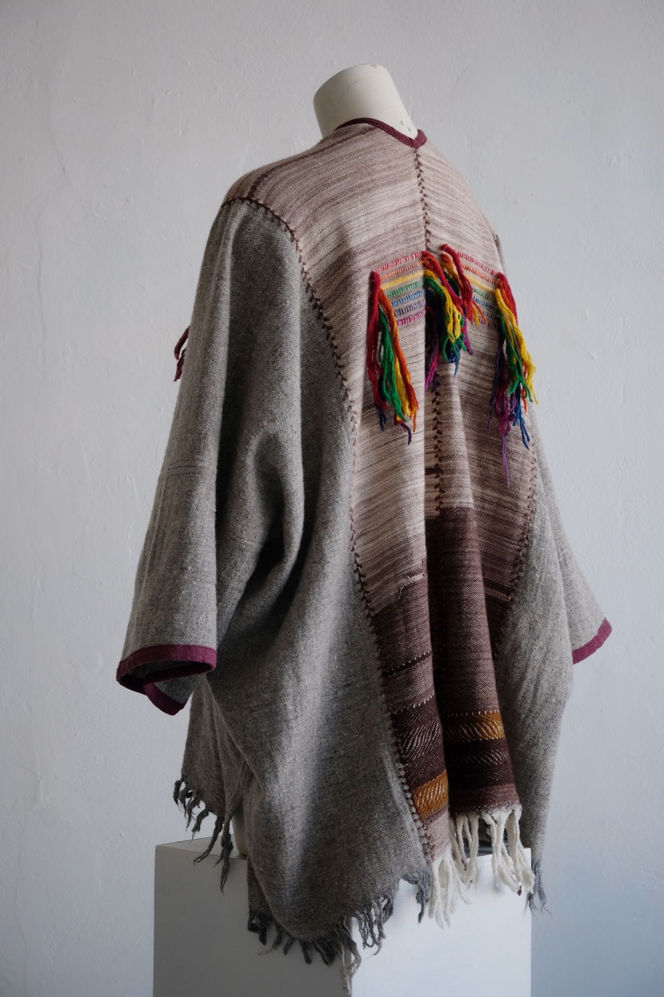 Handwoven, highly textured with fringe cloak that is grey, brown, green and rainbow on a white mannequin against a white wall