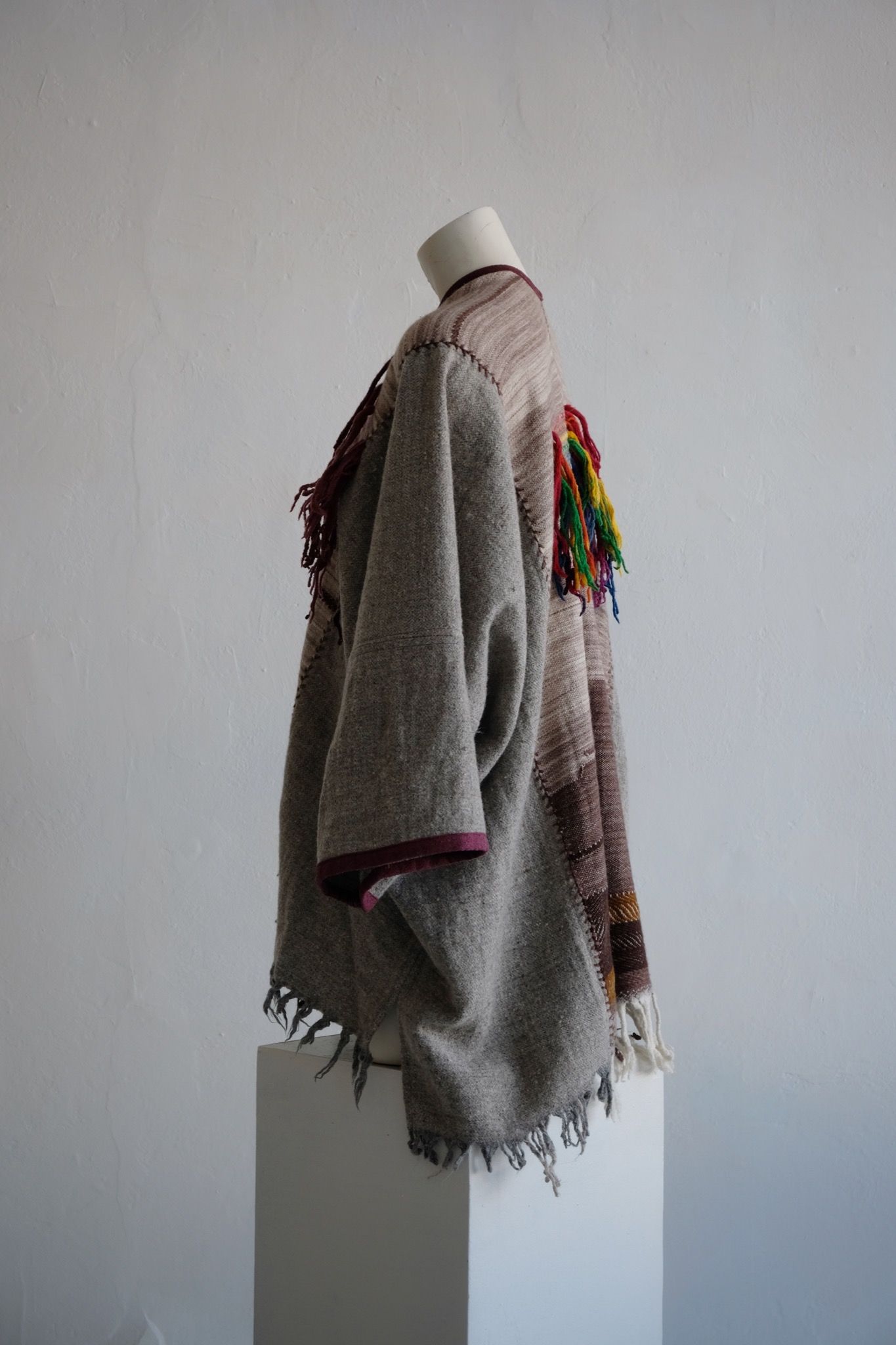 Handwoven, highly textured with fringe cloak that is grey, brown, green and rainbow on a white mannequin against a white wall