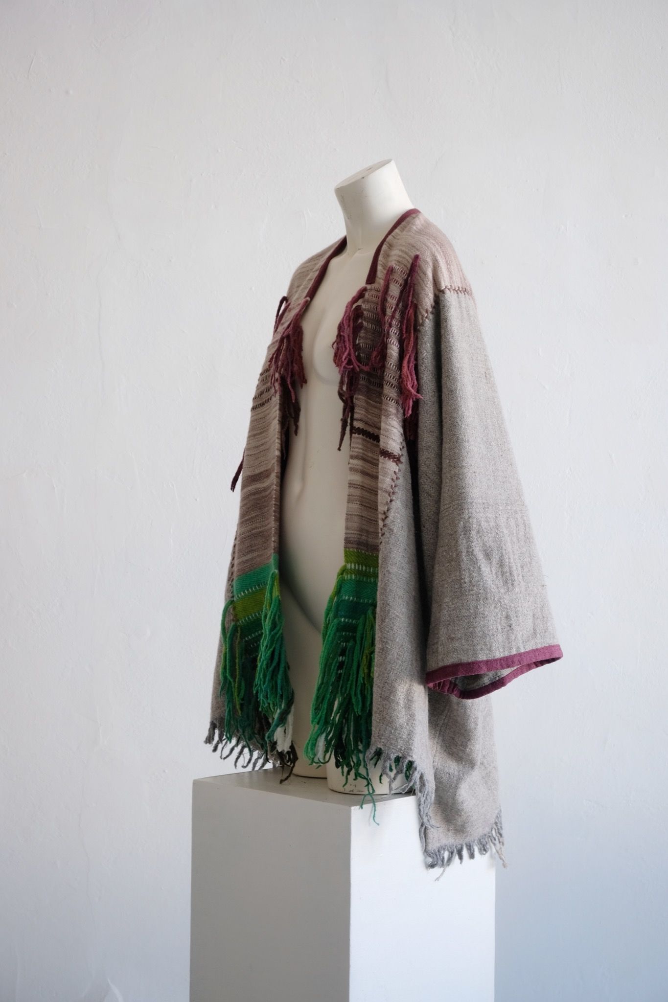 Handwoven, highly textured with fringe cloak that is grey, brown, green and rainbow on a white mannequin against a white wall