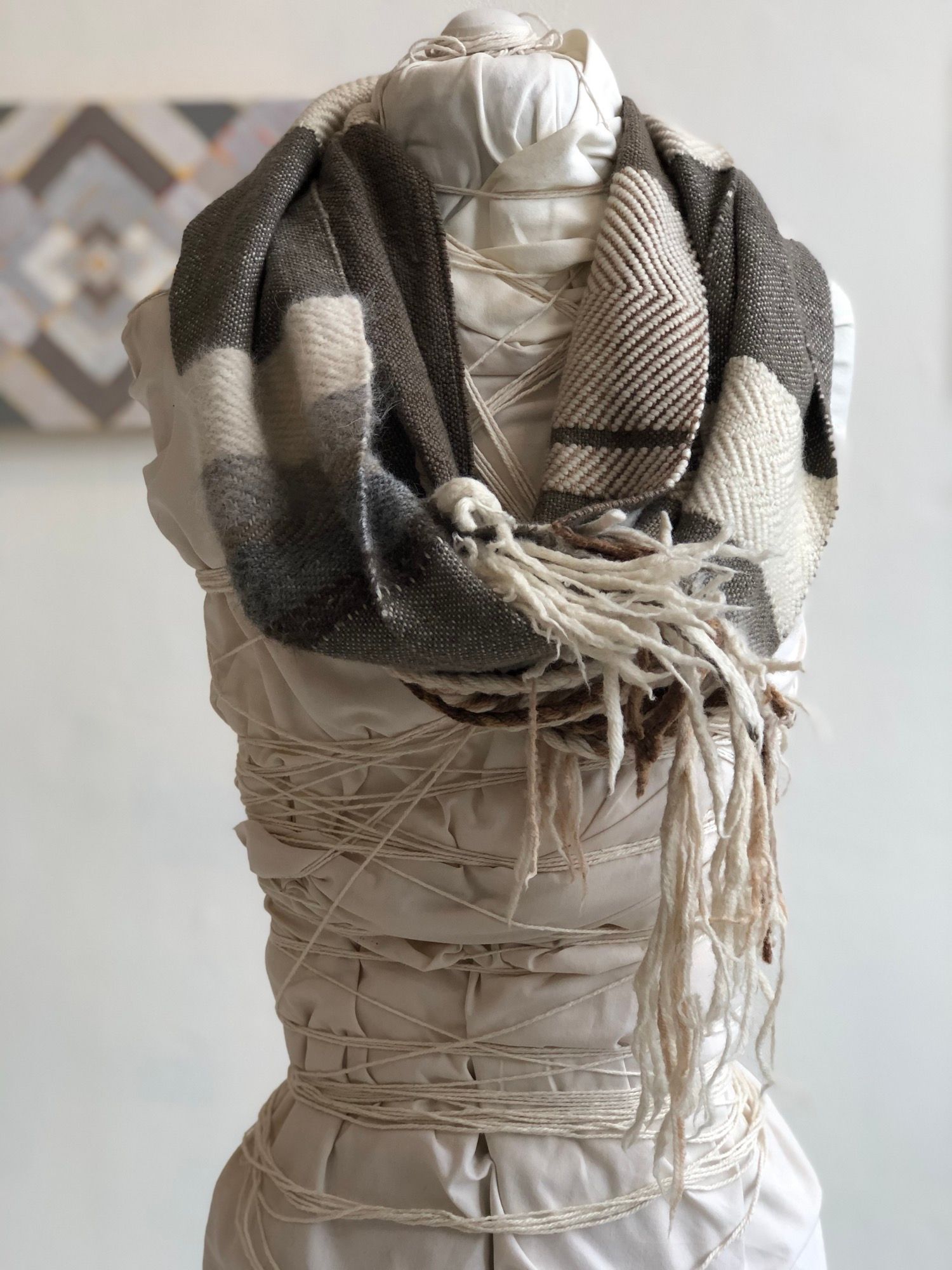 Brown, grey, black and white fringed and braided handwoven cowl scarf on a white mannequin in a gallery