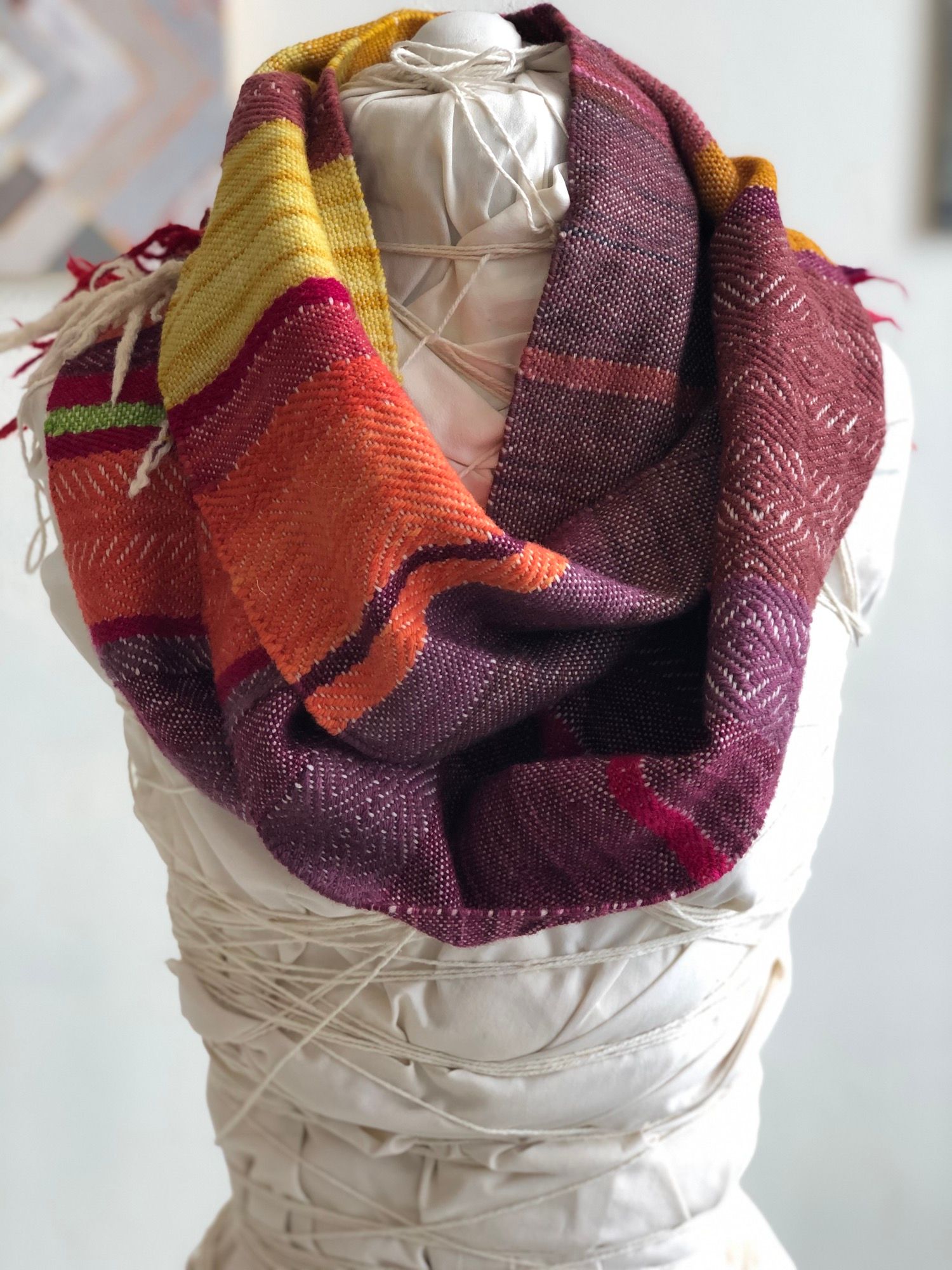 purple, yellow, orange and rainbow handwoven cowl scarf with fringe on a white mannequin in a white walled gallery