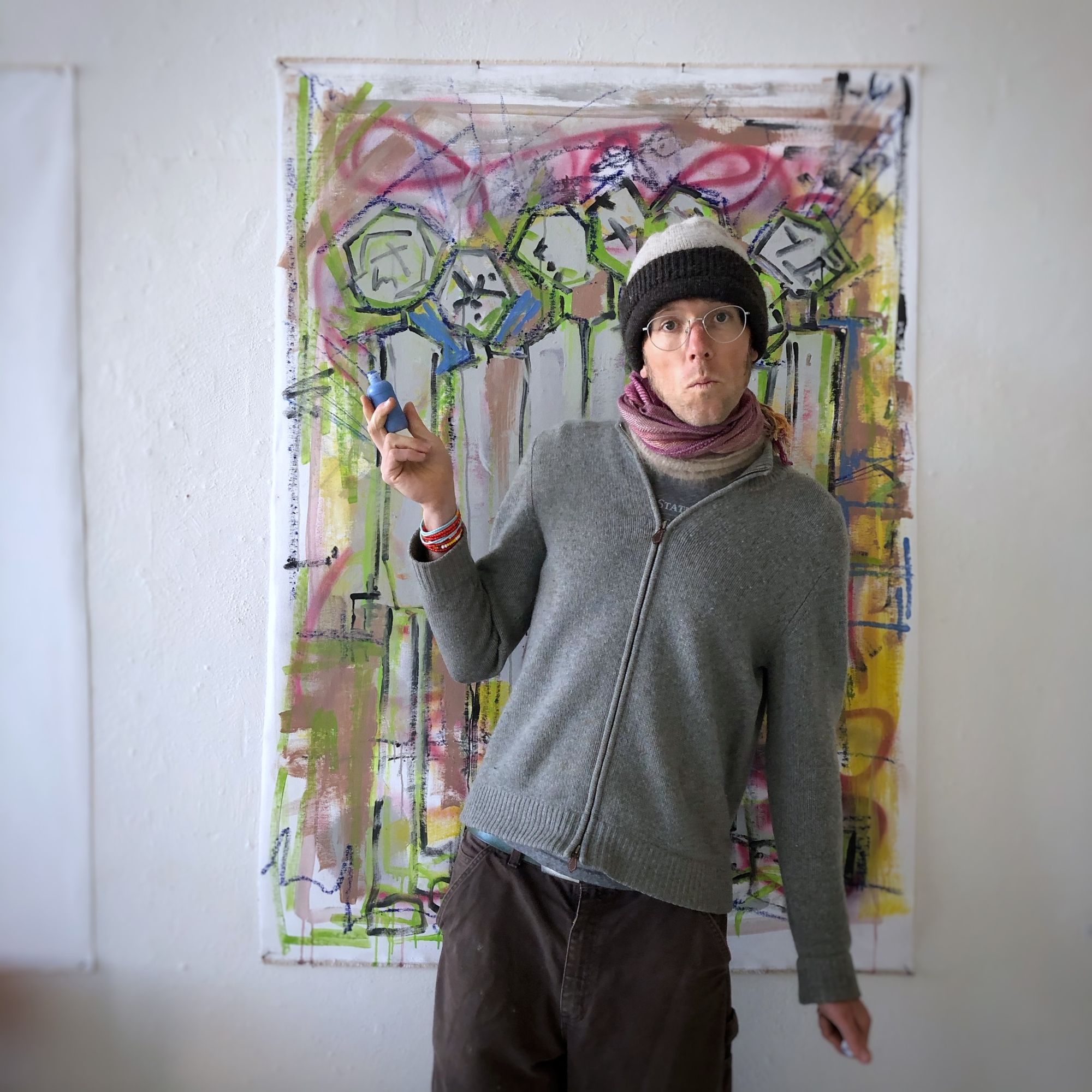 a man in blue sweater and purple scarf holding a paint marker and standing in front of a painting in progress