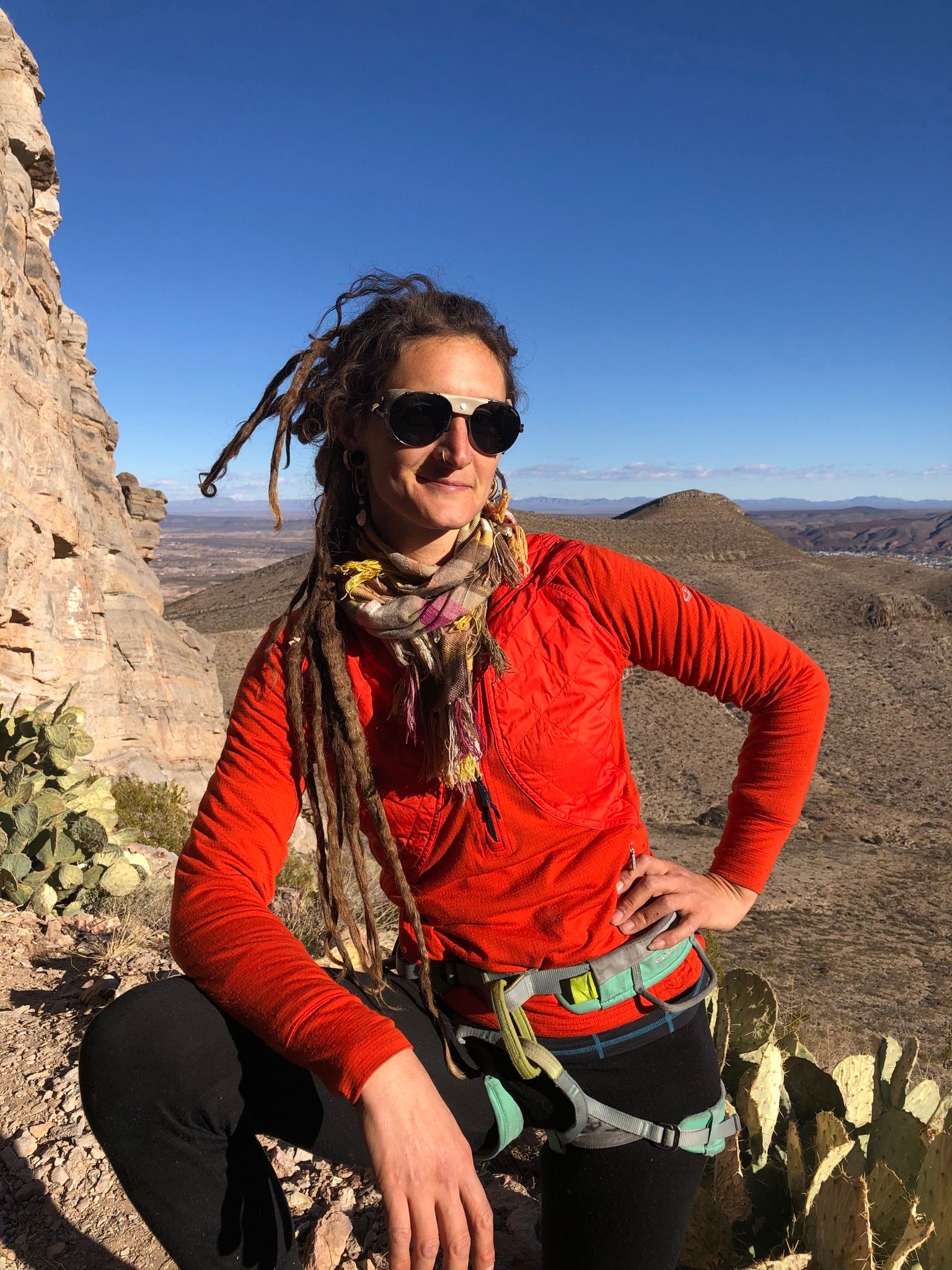 woman in orange shirt, multicolored scarf and sunglasses standing near cactus on a mountain in the desert