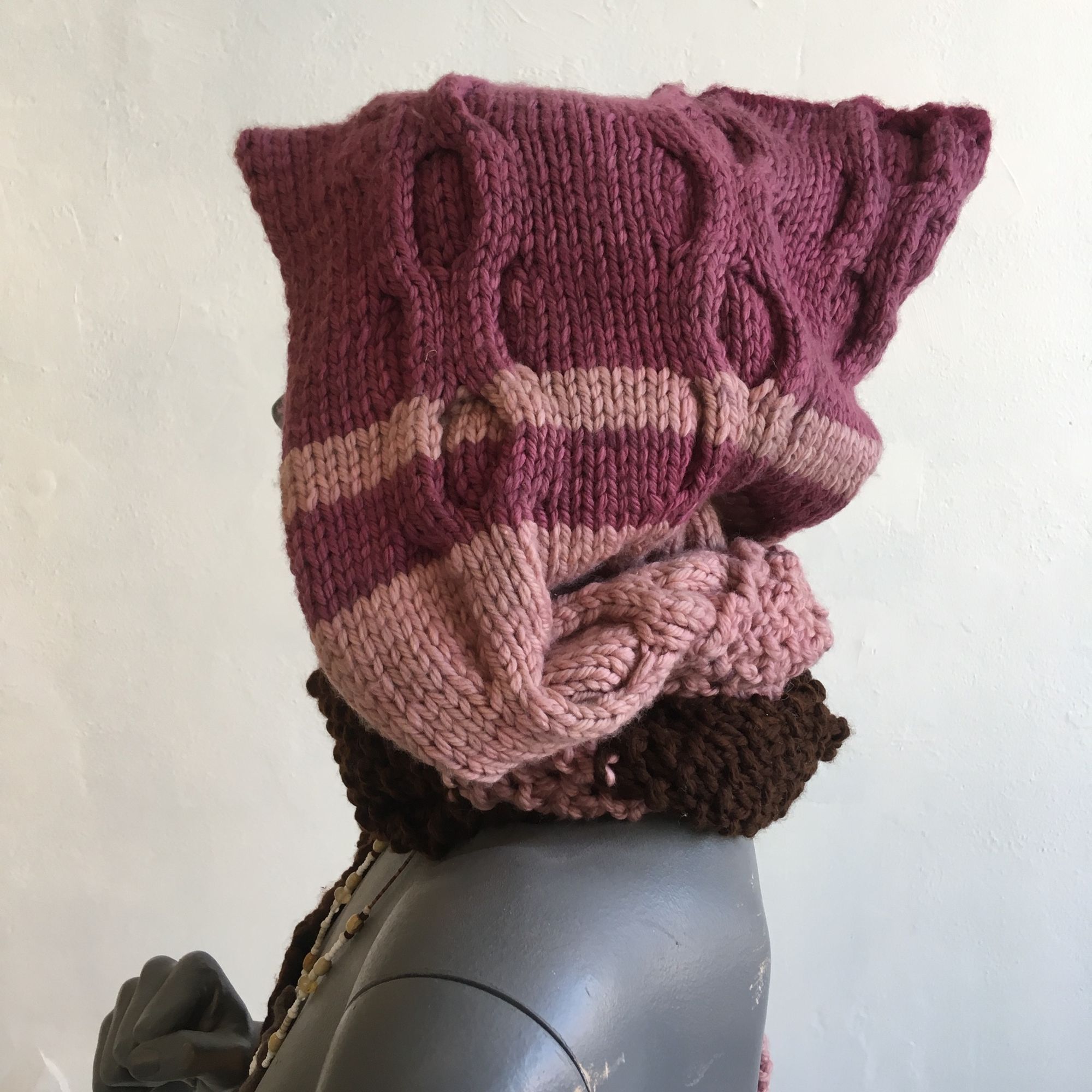 pink and brown cable knit hood-scarf on a grey mannequin against a white wall