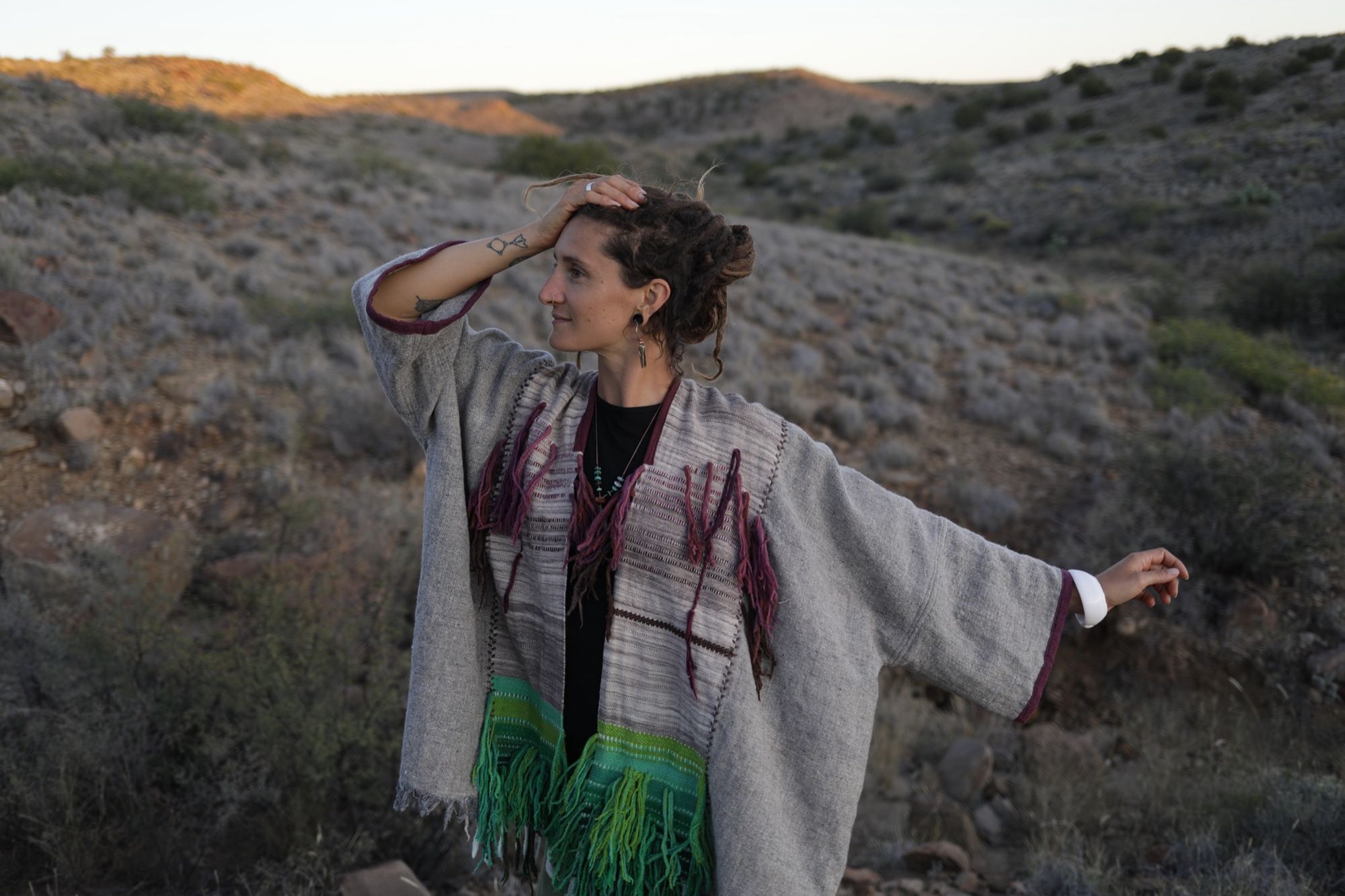 A woman in the desert wearing a Handwoven, highly textured with fringe cloak that is grey, brown, green and rainbow 