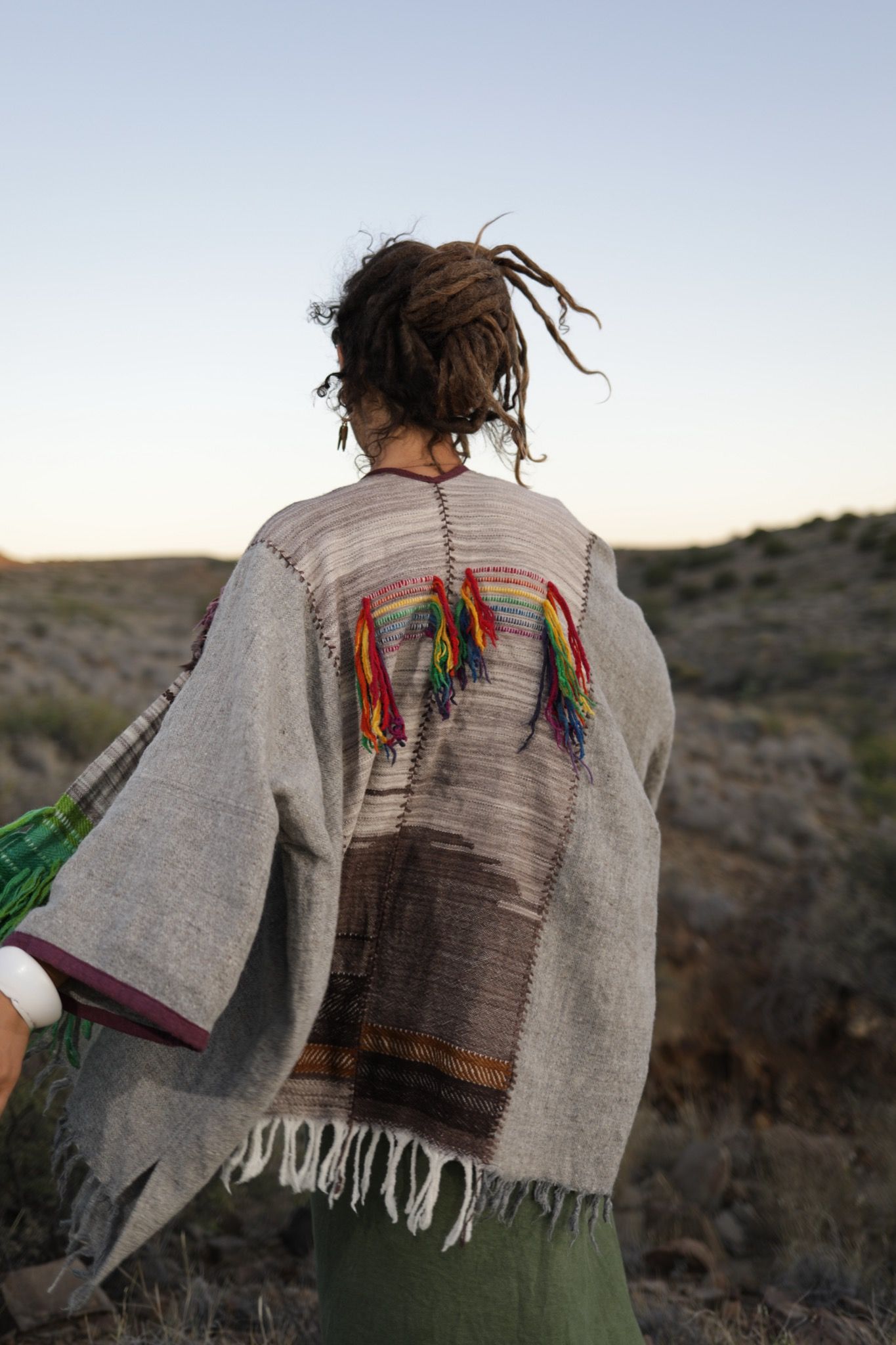 A woman in the desert wearing a Handwoven, highly textured with fringe cloak that is grey, brown, green and rainbow 