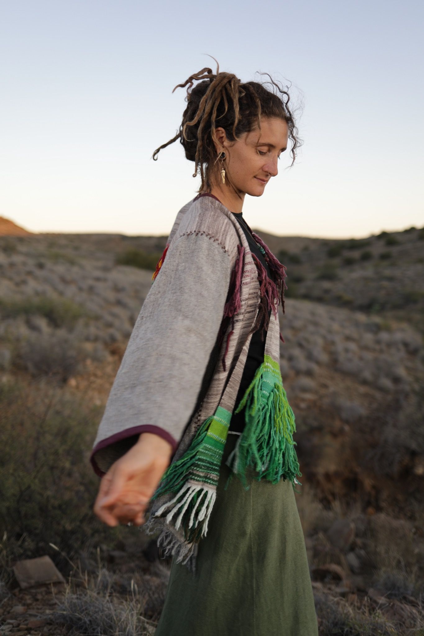 A woman in the desert wearing a Handwoven, highly textured with fringe cloak that is grey, brown, green and rainbow 