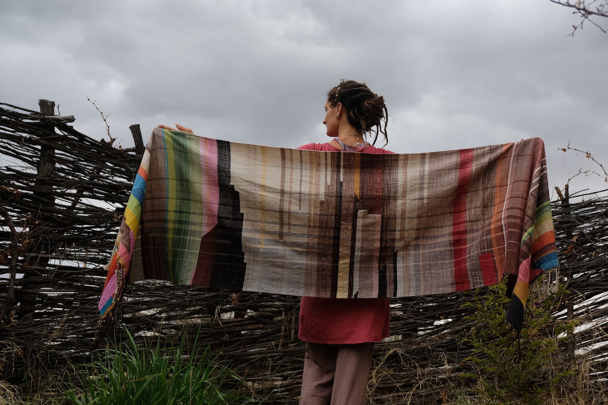 woman wearing a many colored handwoven shawl in front of a stick fence
