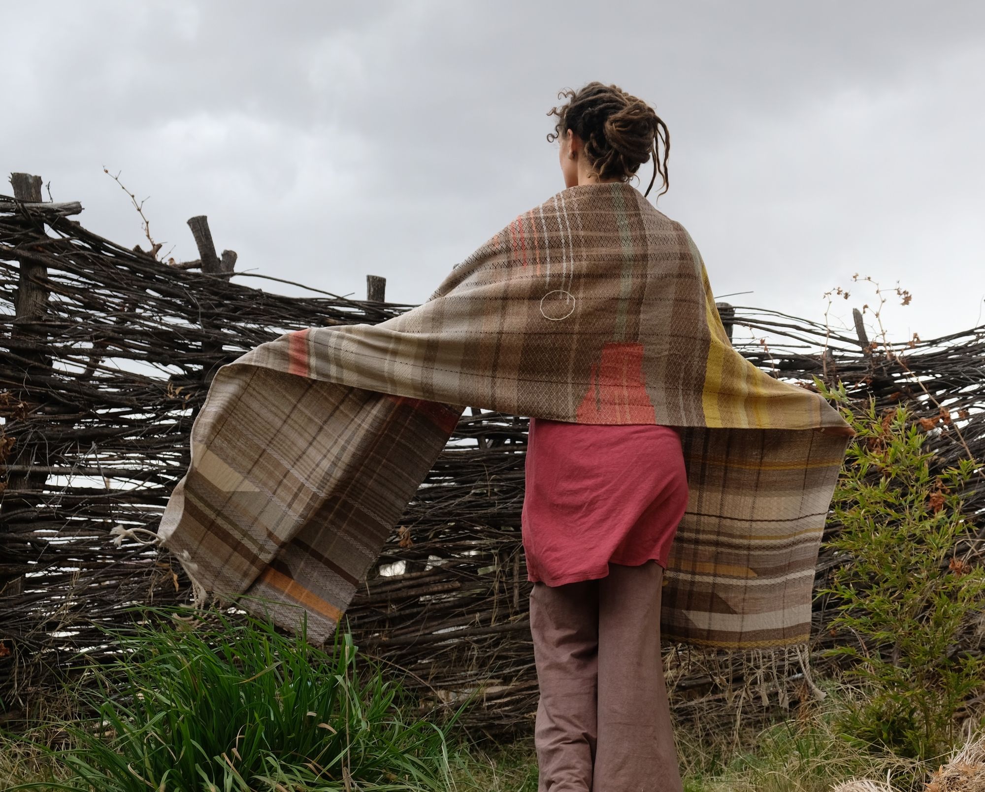 woman wearing a red-pink long-sleeved dress and a handwoven shawl against a stick fence