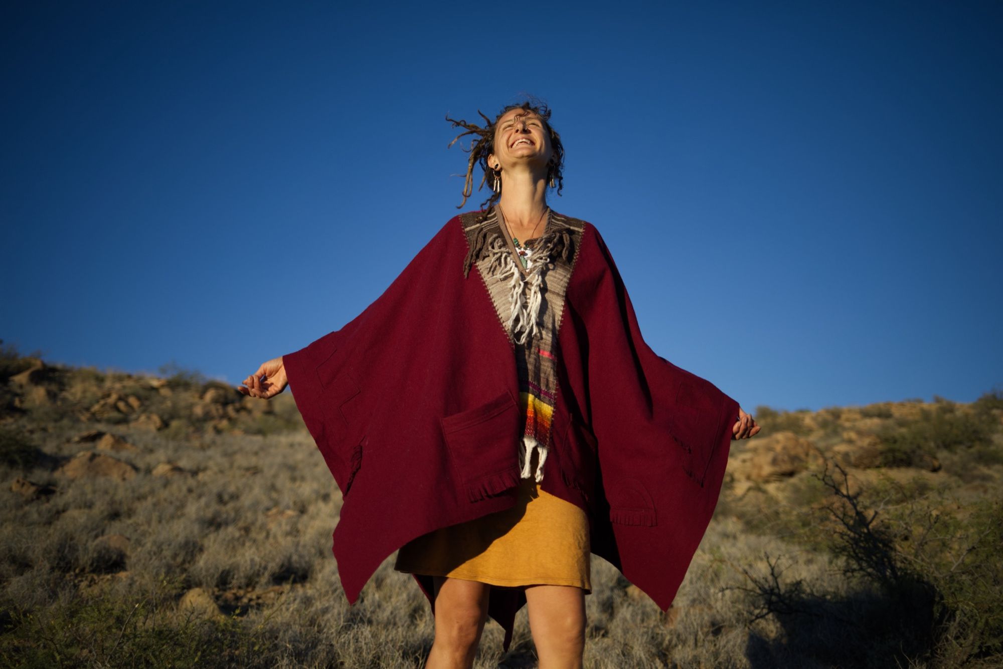 woman wearing a red, brown and rainbow cloak in the desert on a blue sky day