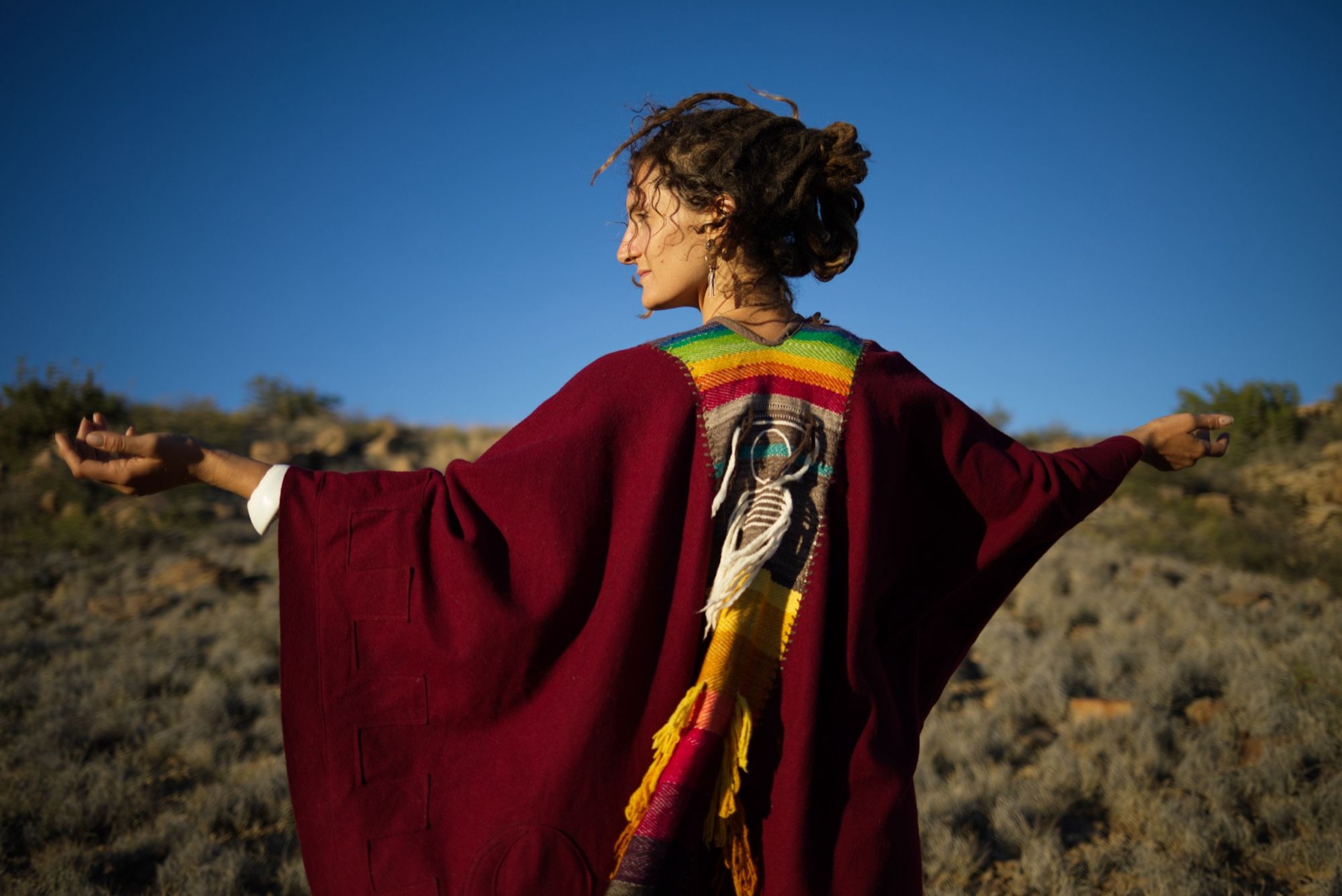 woman wearing a red, brown and rainbow cloak in the desert on a blue sky day