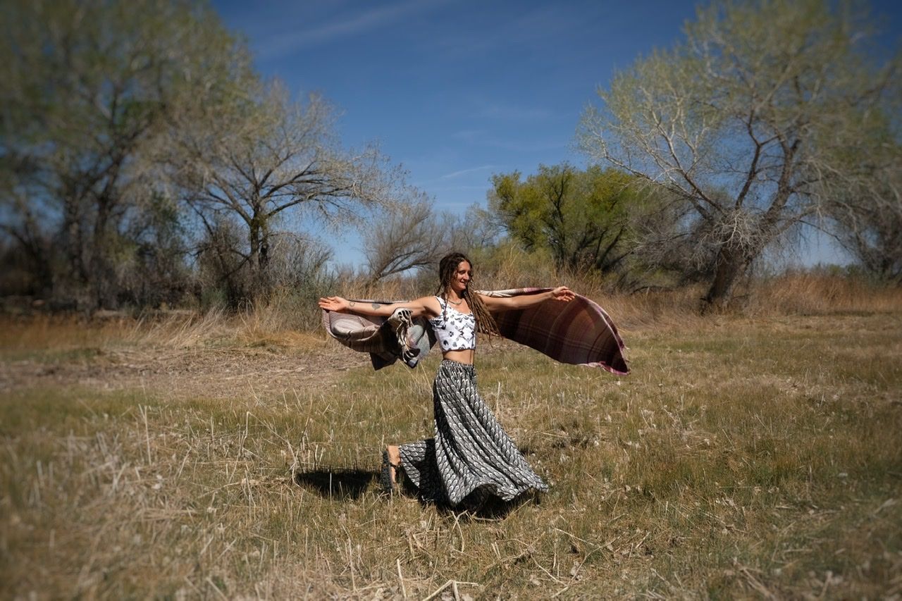 A woman in a black and white skirt and top dances in a grassy field with a pink, blue and grey shawl.  