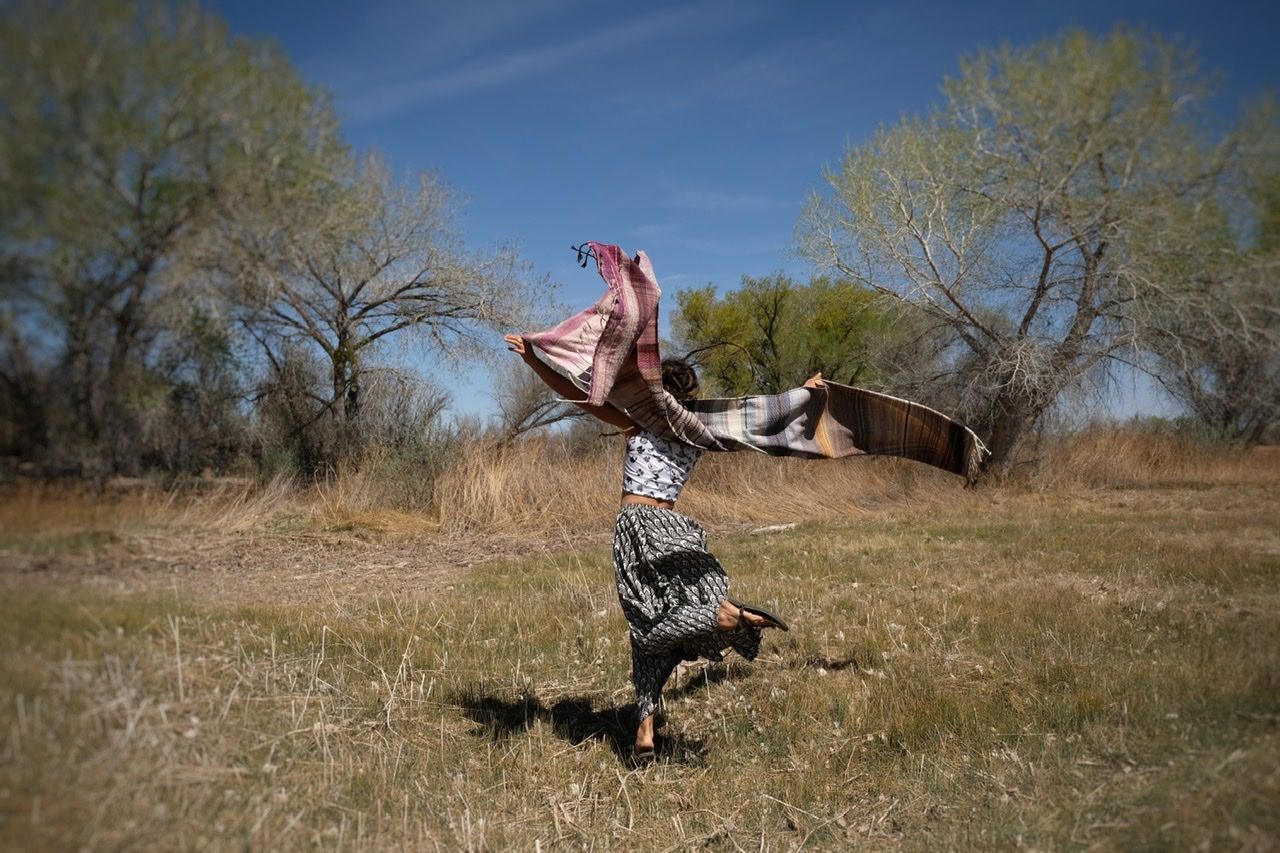 A woman in a black and white skirt and top dances in a grassy field with a pink, blue and grey shawl.  