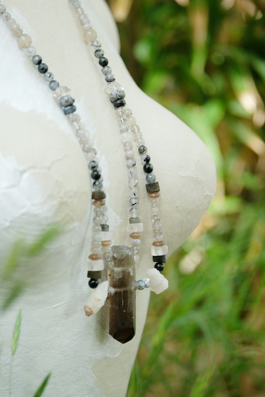 grey smoky quartz and clear crystal stone necklace on a white mannequin surrounded by green plants