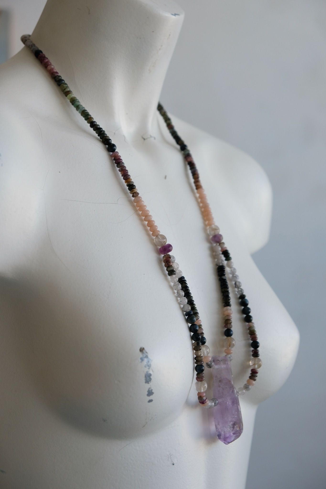 a white mannequin wearing a necklace with a purple amethyst centerpiece and stone beads in subtle shades of pink, grey, black, clear and green