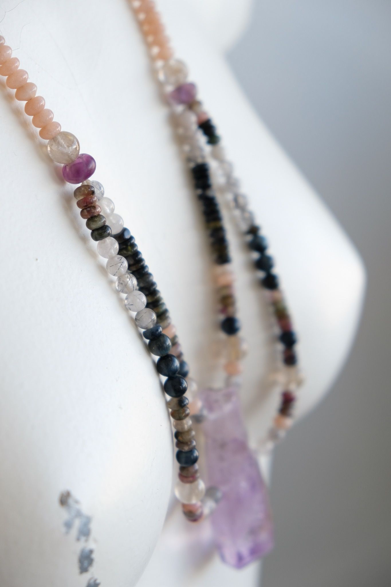 a white mannequin wearing a necklace with a purple amethyst centerpiece and stone beads in pink, grey, black, clear and green