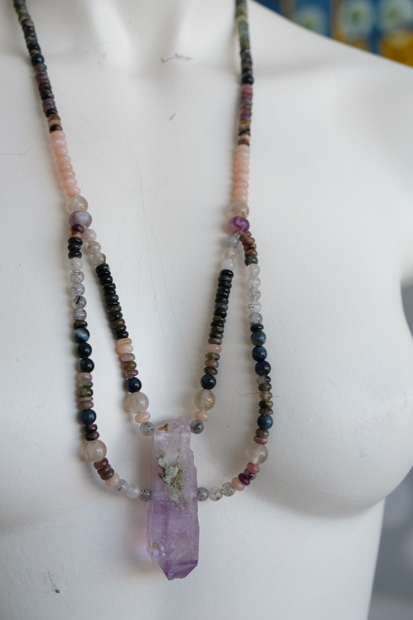 a white mannequin wearing a necklace with a purple amethyst centerpiece and stone beads in pink, grey, black, clear and green