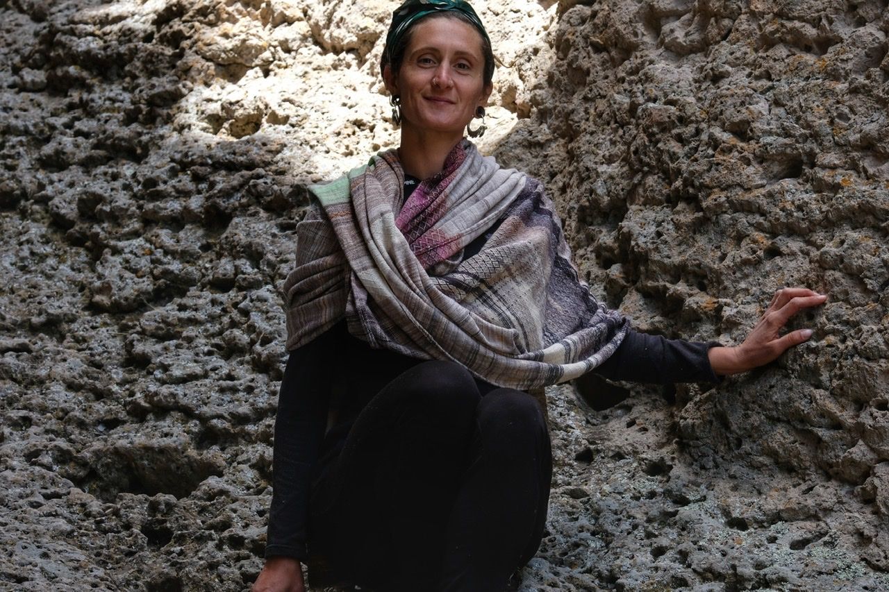 A woman wears a handwoven, diamond pattern shawl in blues, grey, black, brown, pink and green while standing in a stone canyon