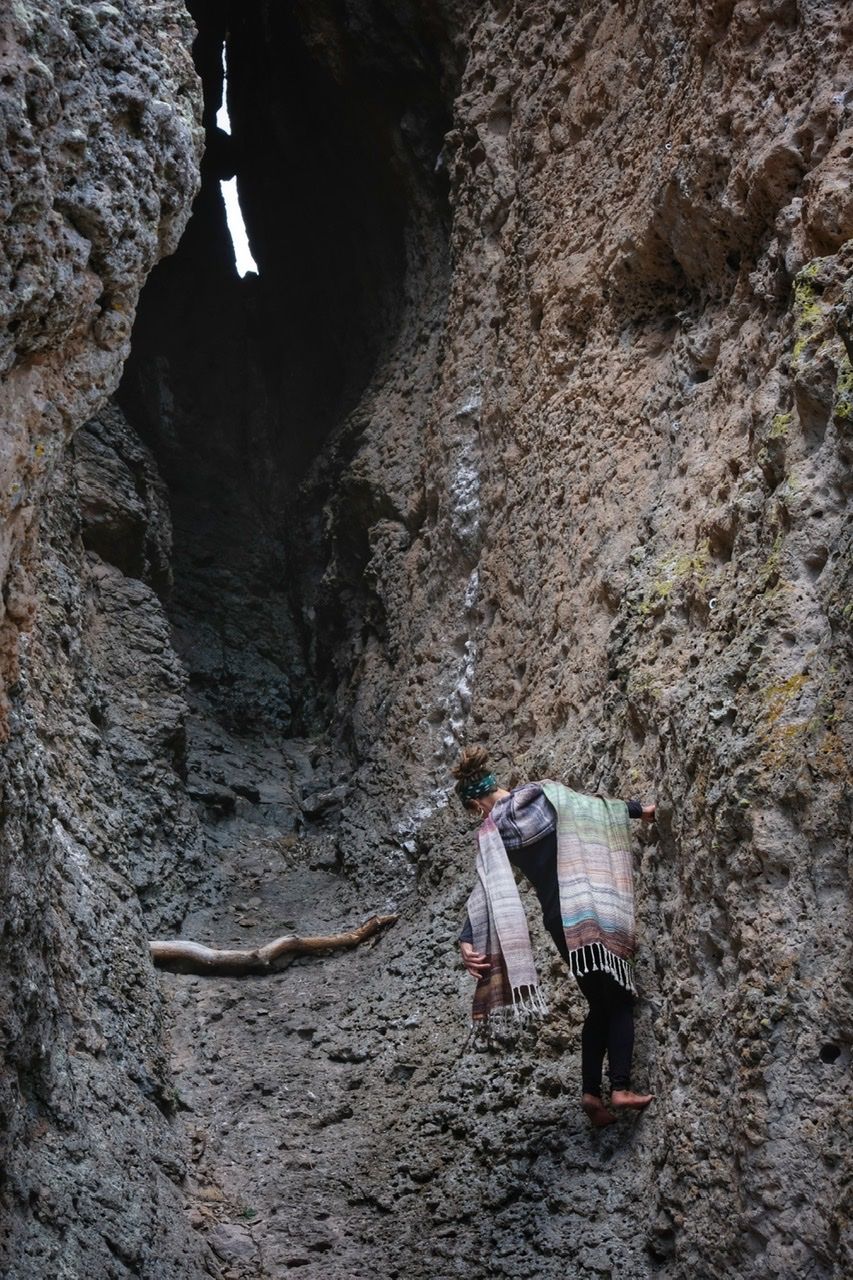 A woman wears a handwoven, diamond pattern shawl in blues, grey, black, brown, pink and green while standing in a stone canyon