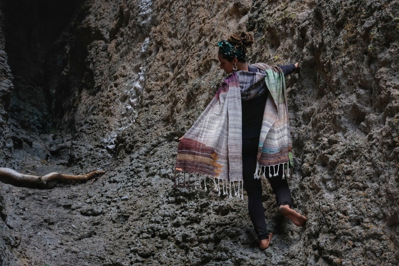 A woman wears a handwoven, diamond pattern shawl in blues, grey, black, brown, pink and green while standing in a stone canyon