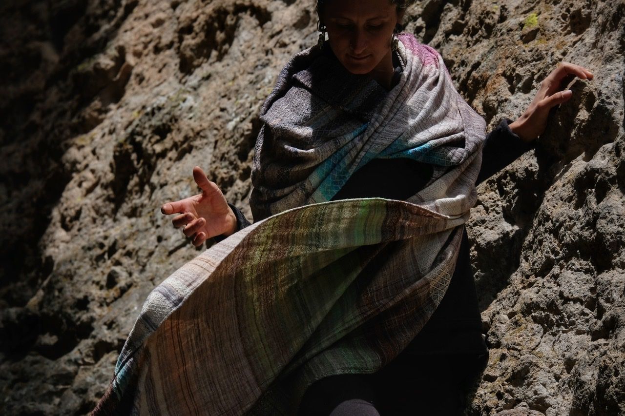 A woman wears a handwoven, diamond pattern shawl in blues, grey, black, brown, pink and green.