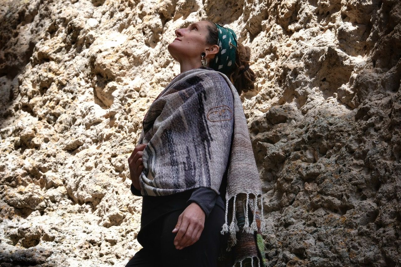 A woman wears a handwoven, diamond pattern shawl in blues, grey, black, brown, pink and green while standing in a stone canyon.